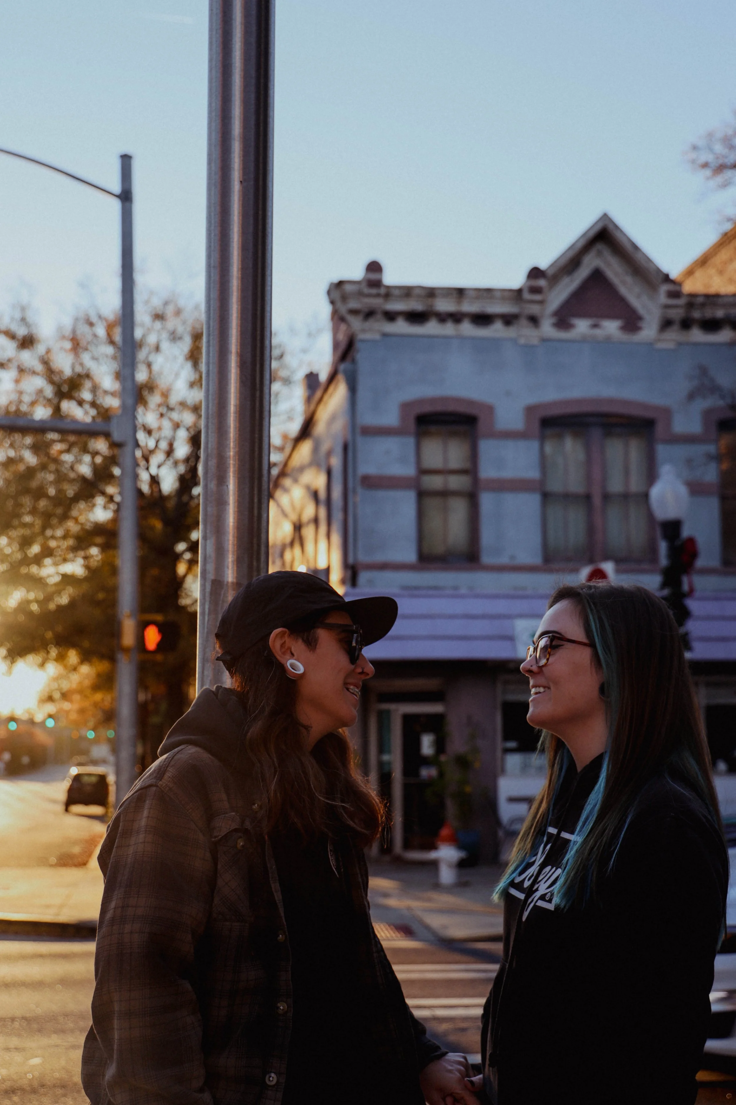 Two women smiling and holding hands on a city street during sunset, with colorful buildings and trees in the background.