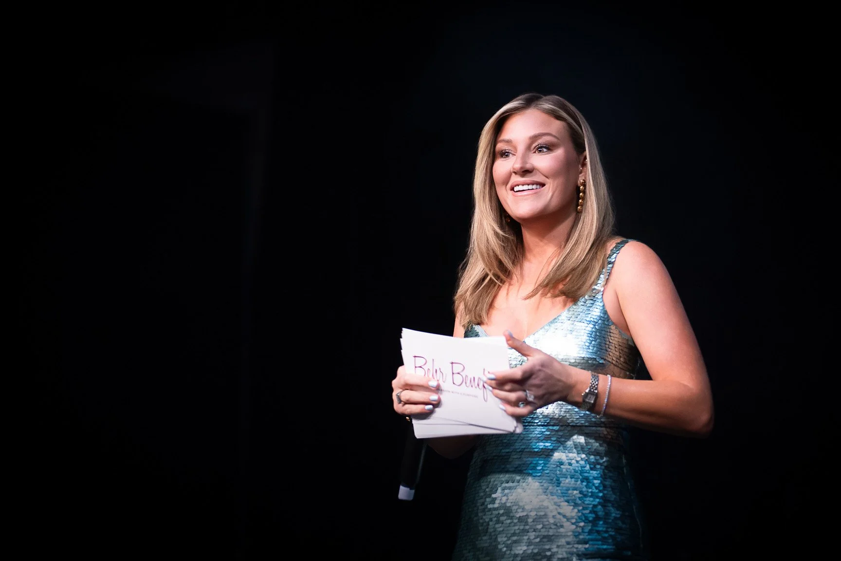 A woman in a shiny blue dress holding a script, smiling, standing against a dark background.