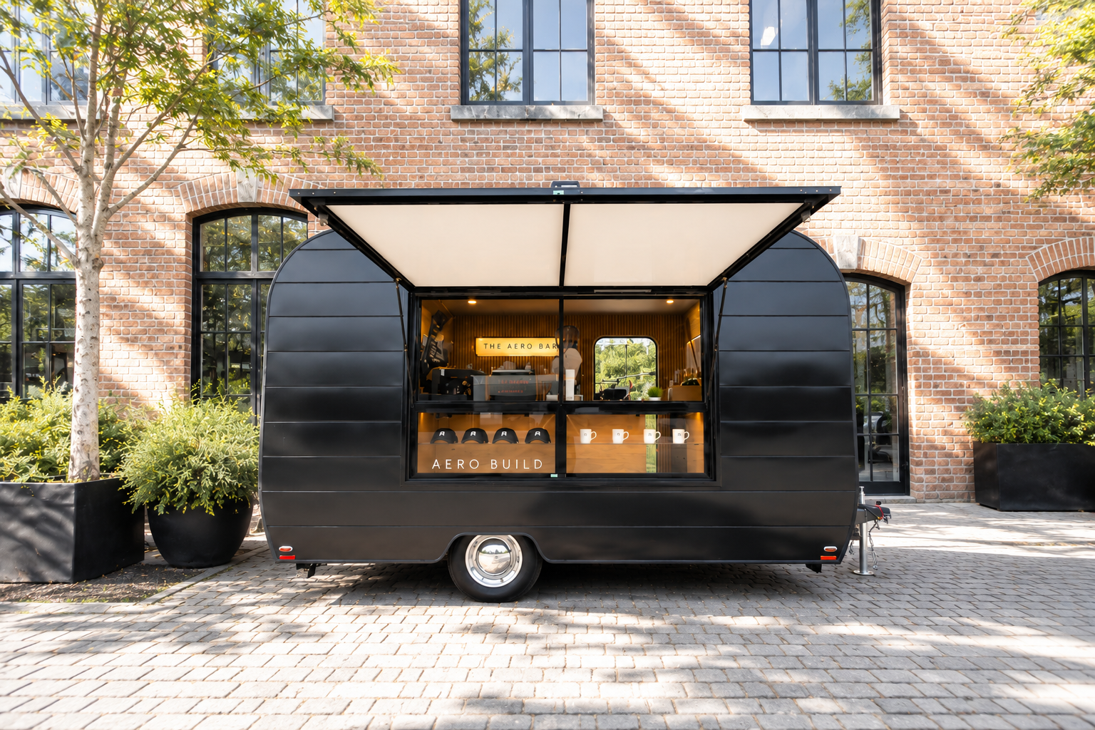 Black mobile coffee shop trailer parked on cobblestone street in front of brick building with large windows and potted plants on either side. The trailer has an open serving window with a person inside and a sign that reads 'The Aero Bar' and 'Aero Build' with coffee mugs on display.