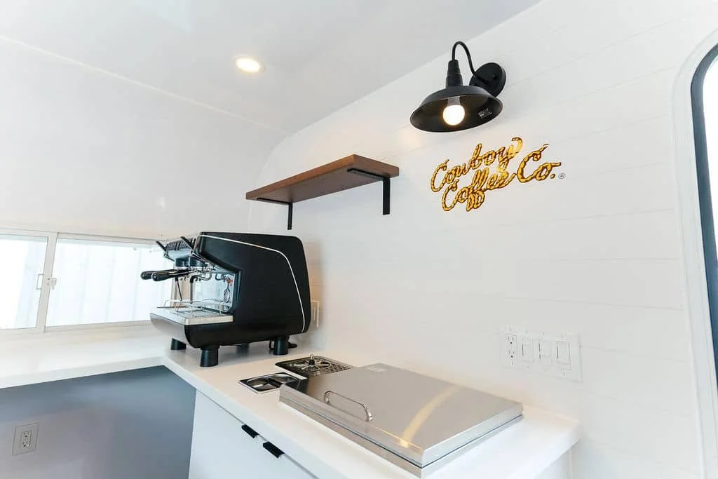 A small, modern kitchen corner with a black espresso machine on a white countertop, a stainless steel cover, a wall-mounted light, a small wooden shelf, and a gold sign reading "Cowboy Coffee Co."