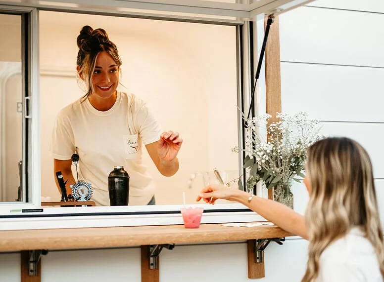 a girl hands another girl a pink drink in a cup with a straw.