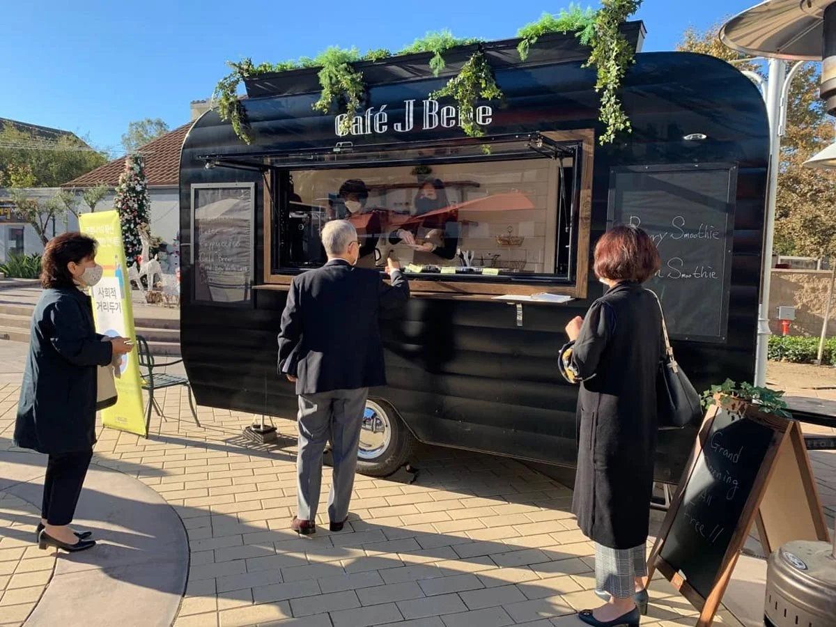 Black food truck with a serving window, decorated with greenery, selling smoothies and sandwiches. Three customers wearing masks are standing in line, and two staff members are inside preparing orders. The scene is outdoors on a sunny day.
