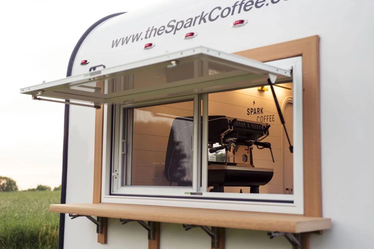 A coffee stand inside a mobile trailer with the website theSparkCoffee.com displayed on the outside, a window serving coffee, with the interior featuring a coffee machine, and a sign reading 'SPARK COFFEE' inside.