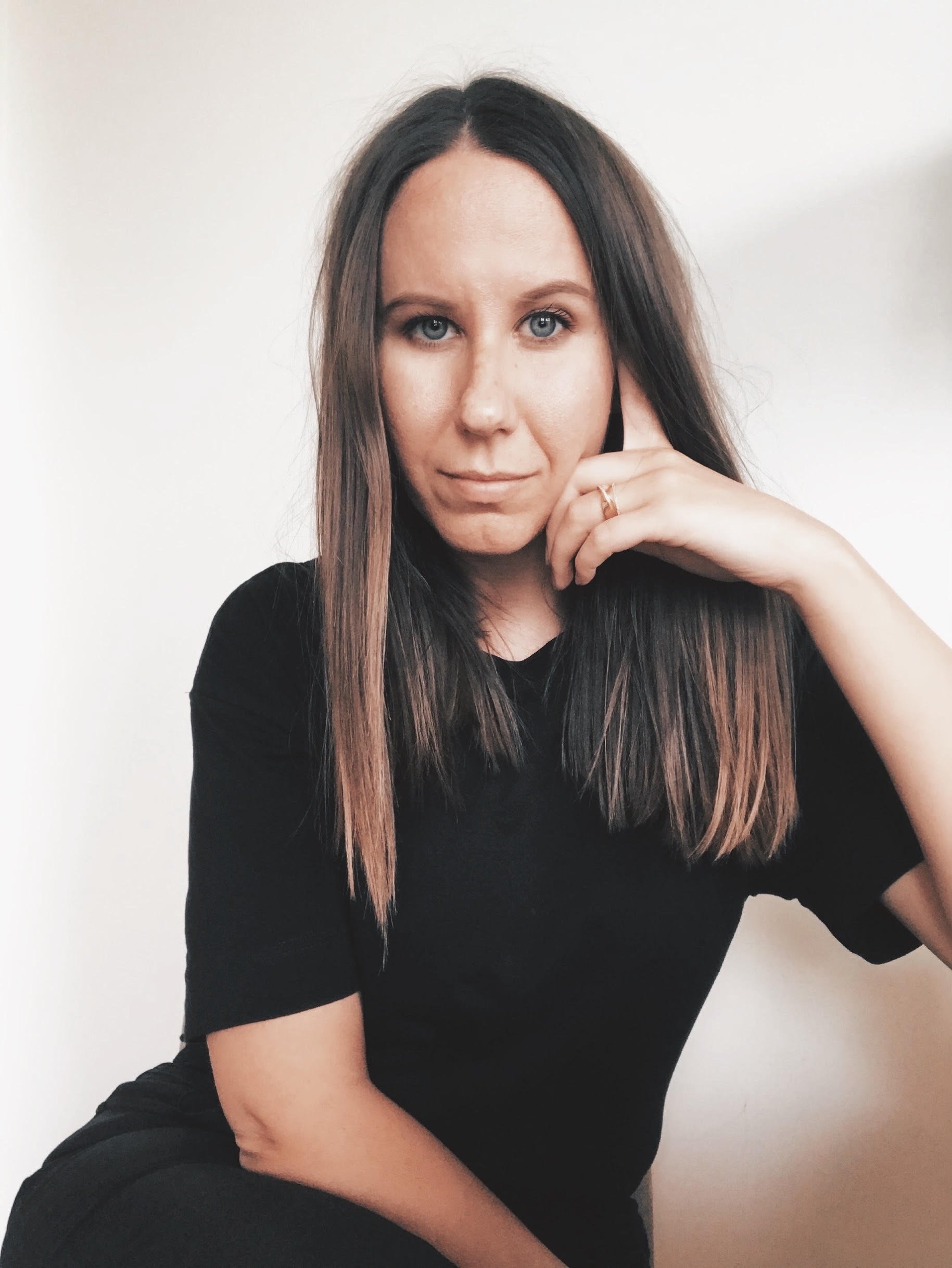A woman with long brown hair and blue eyes wearing a black shirt, sitting with her hand resting on her cheek, against a plain white background.