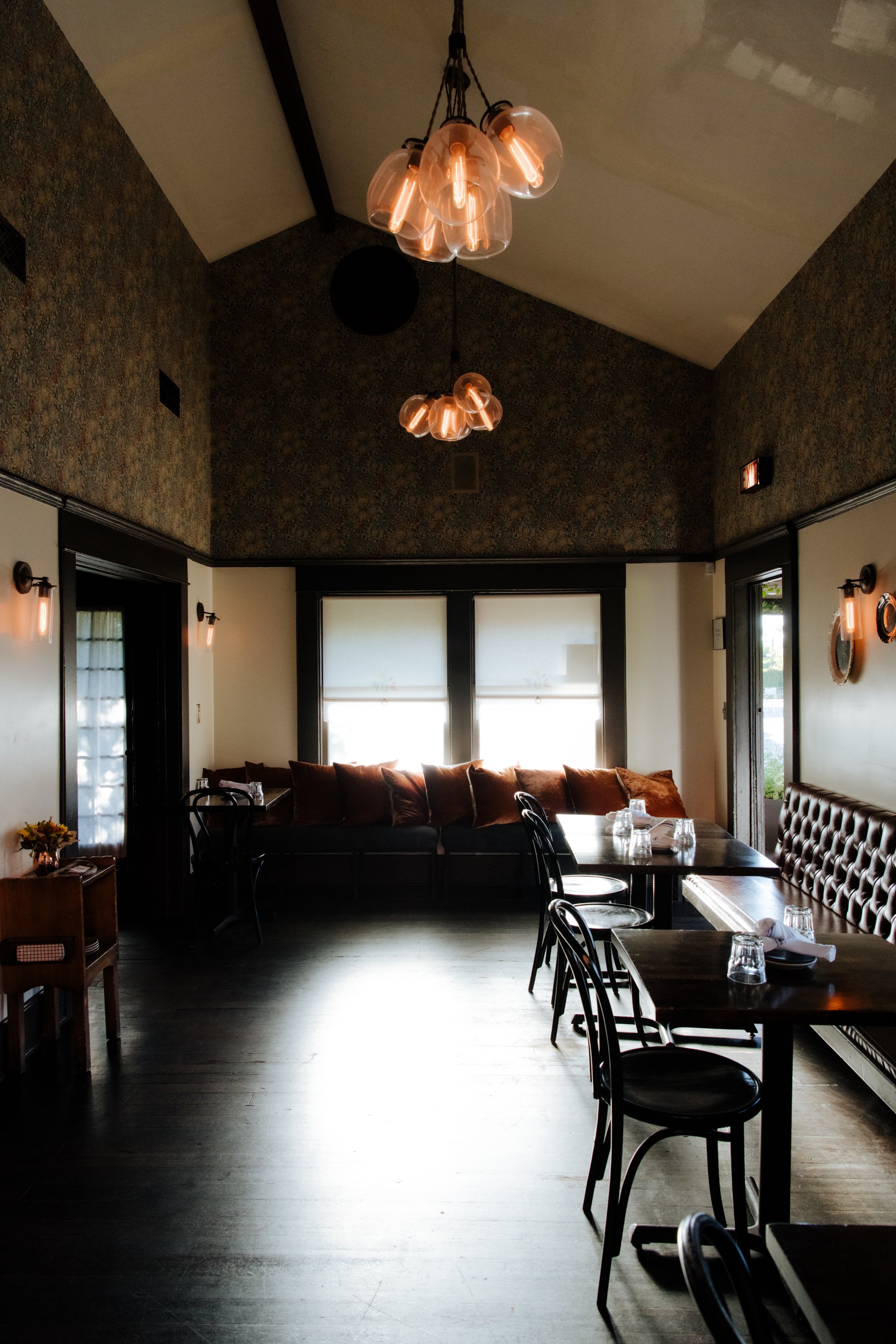 Interior of a cozy café with a high ceiling, patterned wallpaper, modern hanging lights, and a mix of seating including sofas and chairs beside large windows letting in natural light.