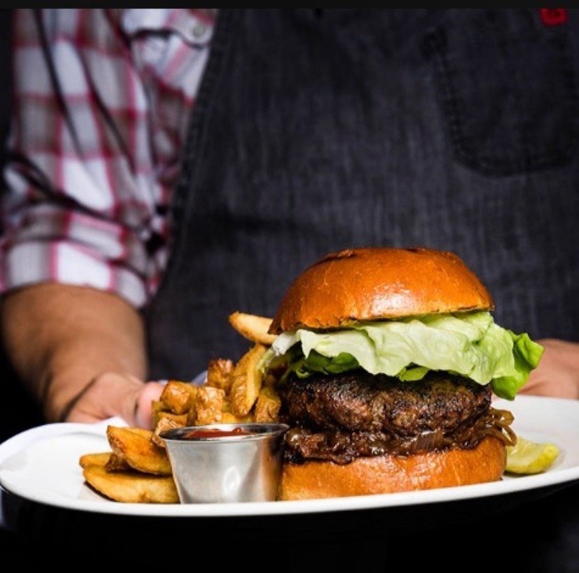 A person in a denim apron presents a plate holding a large burger with lettuce and a side of fries and a dipping sauce.