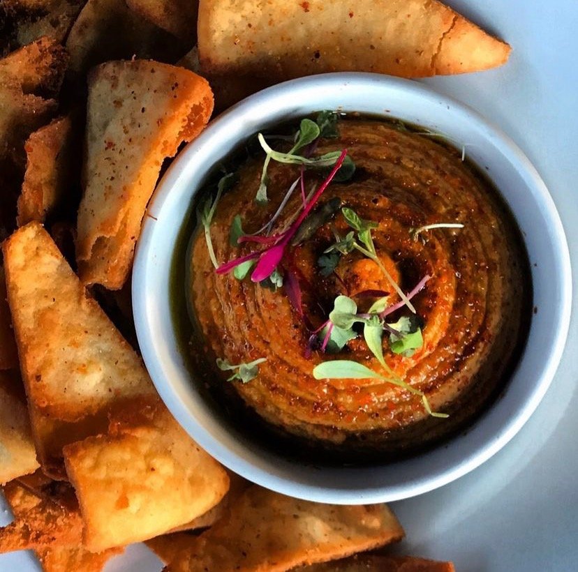 A bowl of roasted red pepper hummus garnished with microgreens, surrounded by triangular pita chips, served on a white plate.