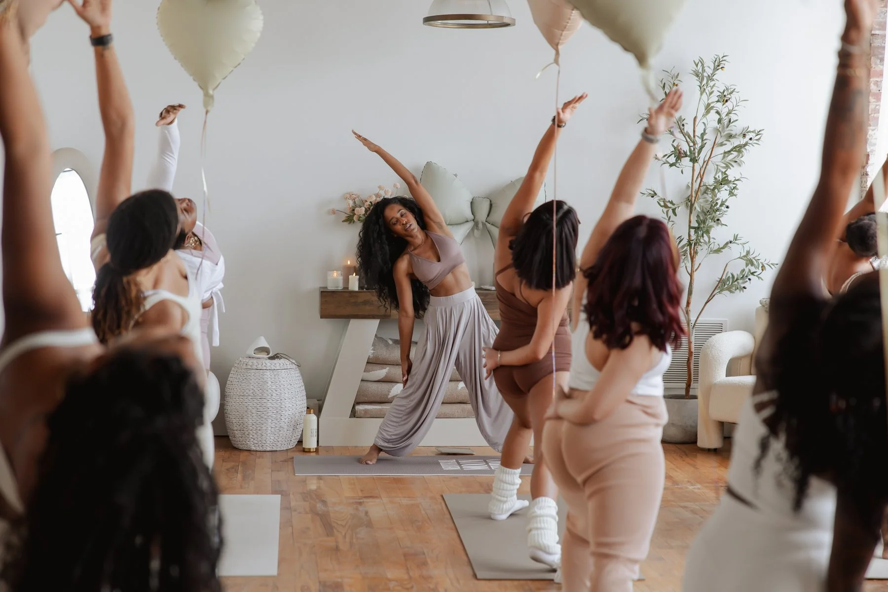 A group of women participating in a yoga class in a bright, cozy living room. The instructor is leading the class, demonstrating a stretching pose. The women are standing on yoga mats, wearing comfortable workout clothes, and appear focused and relax
