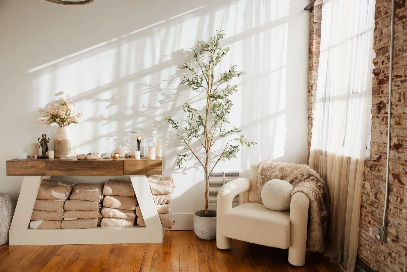 Cozy interior with a white armchair, a plush cushion, a fur throw, a tall potted plant, and a wooden console table with candles, a vase, and decorative items, illuminated by natural sunlight through a sheer curtain and brick wall.