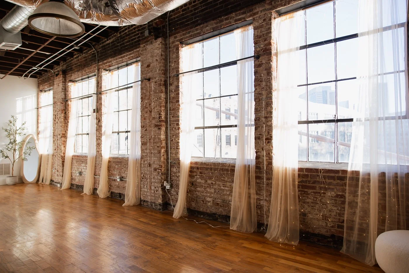Empty room with brick walls, large industrial-style windows with sheer curtains, wooden floor, and a potted plant.