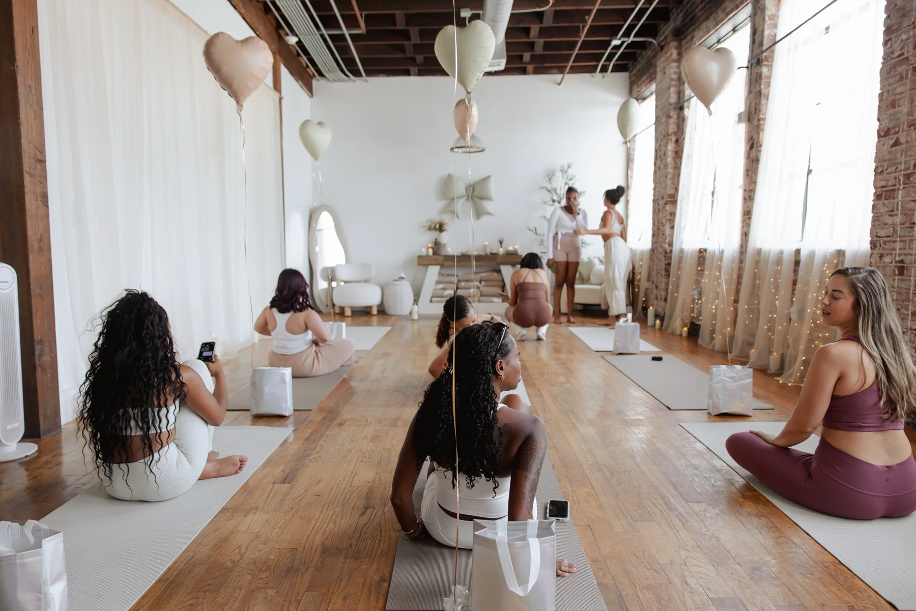 Women participating in a yoga or meditation class in a spacious, decorated room with white and brick walls, large windows with curtains, and heart-shaped balloons hanging from the ceiling.