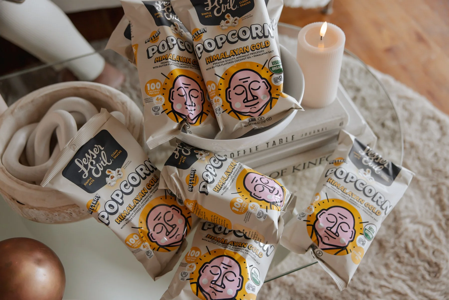 Several bags of Patty's Evil popcorn in Himalayan Gold flavor on a glass table, with a white candle and a white tray underneath, and a ceramic bowl with white rings, with a wooden floor and a small part of a person's foot and leg in background.