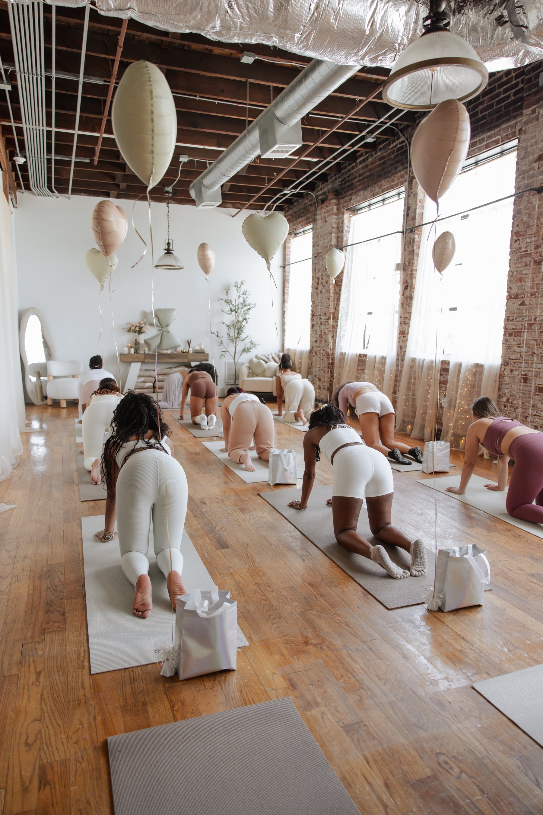 People participating in a prenatal yoga class in a bright, spacious room with large windows, exposed brick walls, and wooden floors. They are practicing yoga on mats, with some in a kneeling or forward bend position, and include individuals with vari