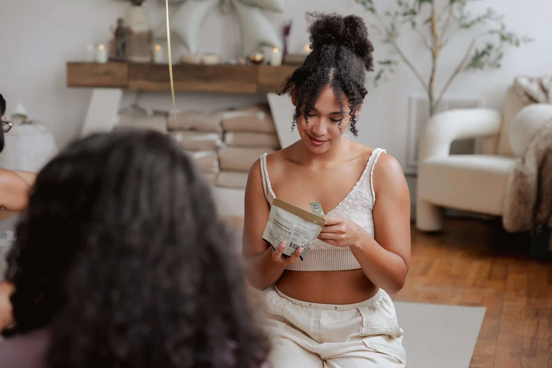 A young woman with curly dark hair tied in a high puff sits on the floor, smiling and looking at a snack package she is holding, in a cozy living room with beige furniture and a wooden floor.