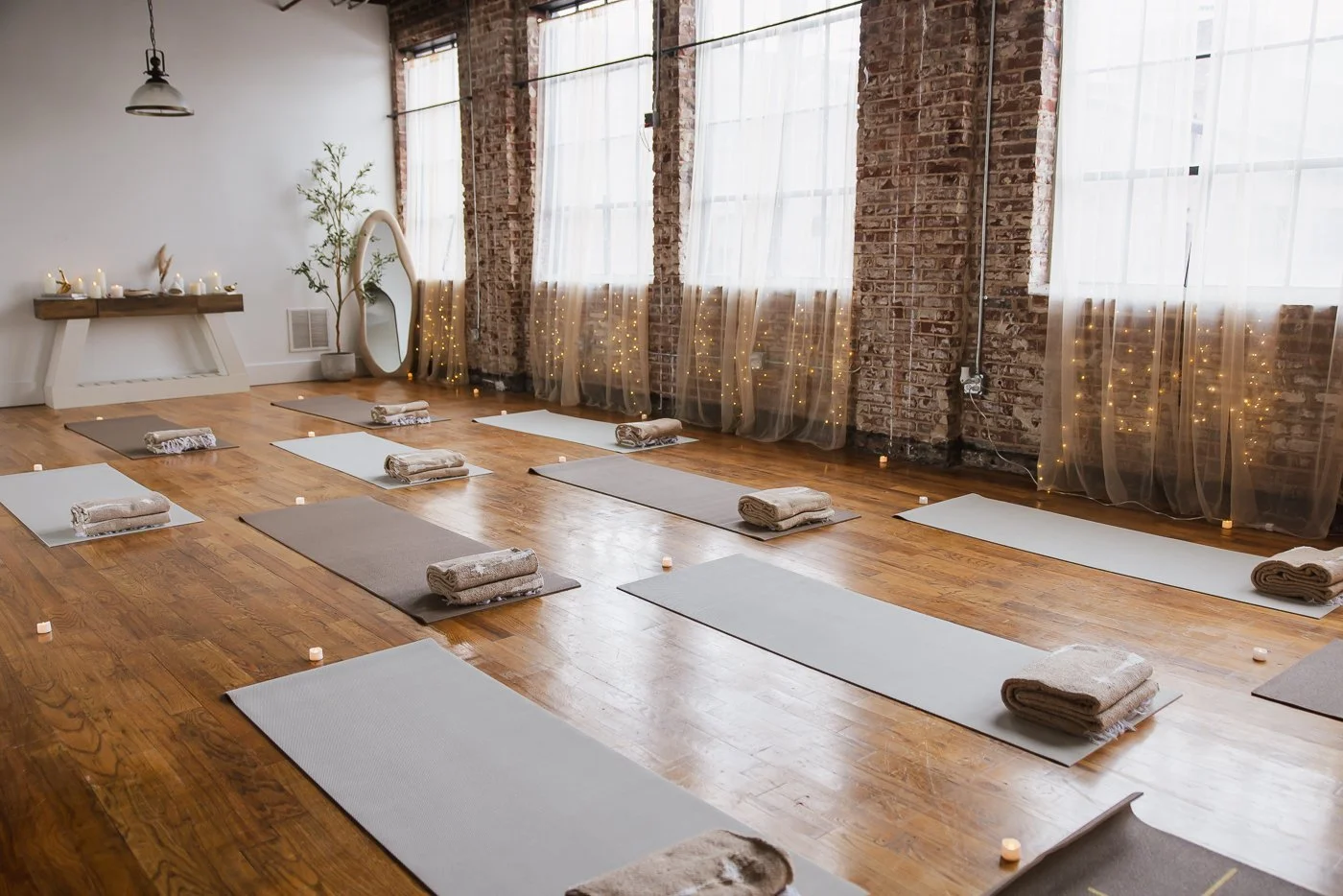 A yoga or meditation studio with hardwood floors, white and gray mats, rolled towels, small candles, string lights behind sheer curtains, and a rustic shelf against a white wall with candles and a decorative mirror.