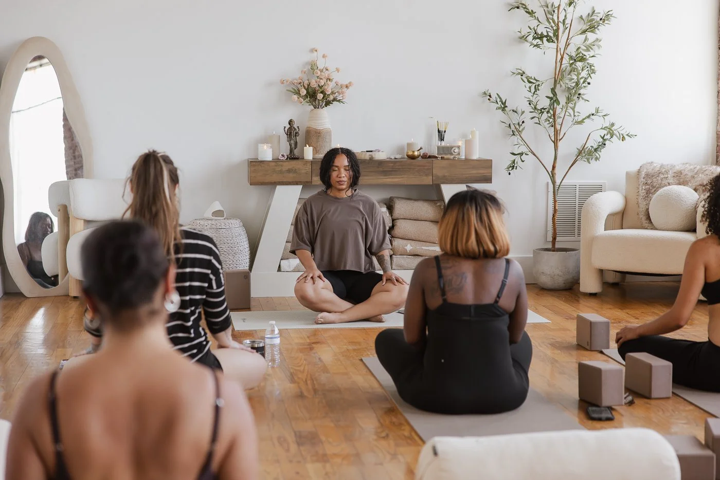 Group of women participating in a yoga or meditation class in a cozy, well-lit living room with wooden floors, potted plants, and minimal decor.