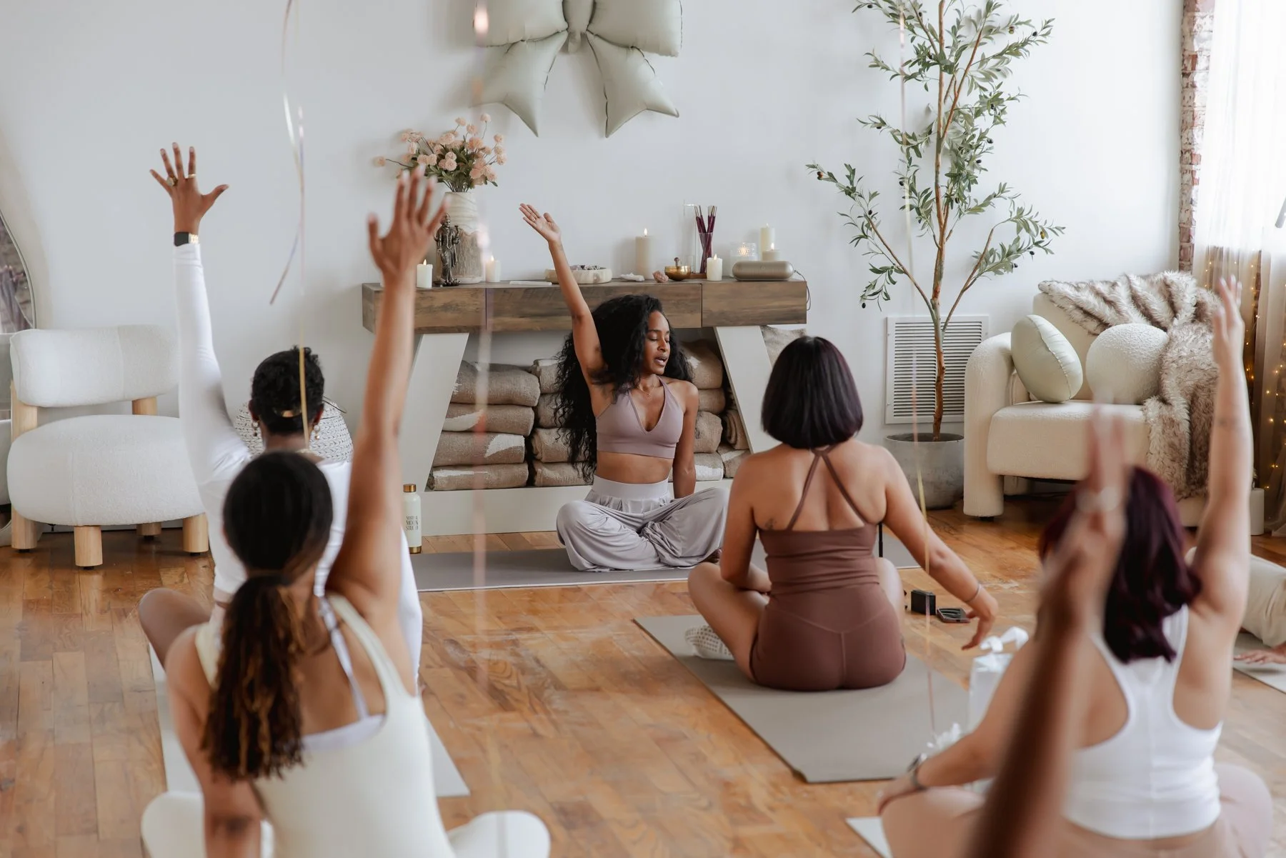 A group of people participating in a yoga or meditation class in a cozy, well-decorated room with a woman leading the session. Some participants are raising their hands while sitting on yoga mats, engaging in the activity.