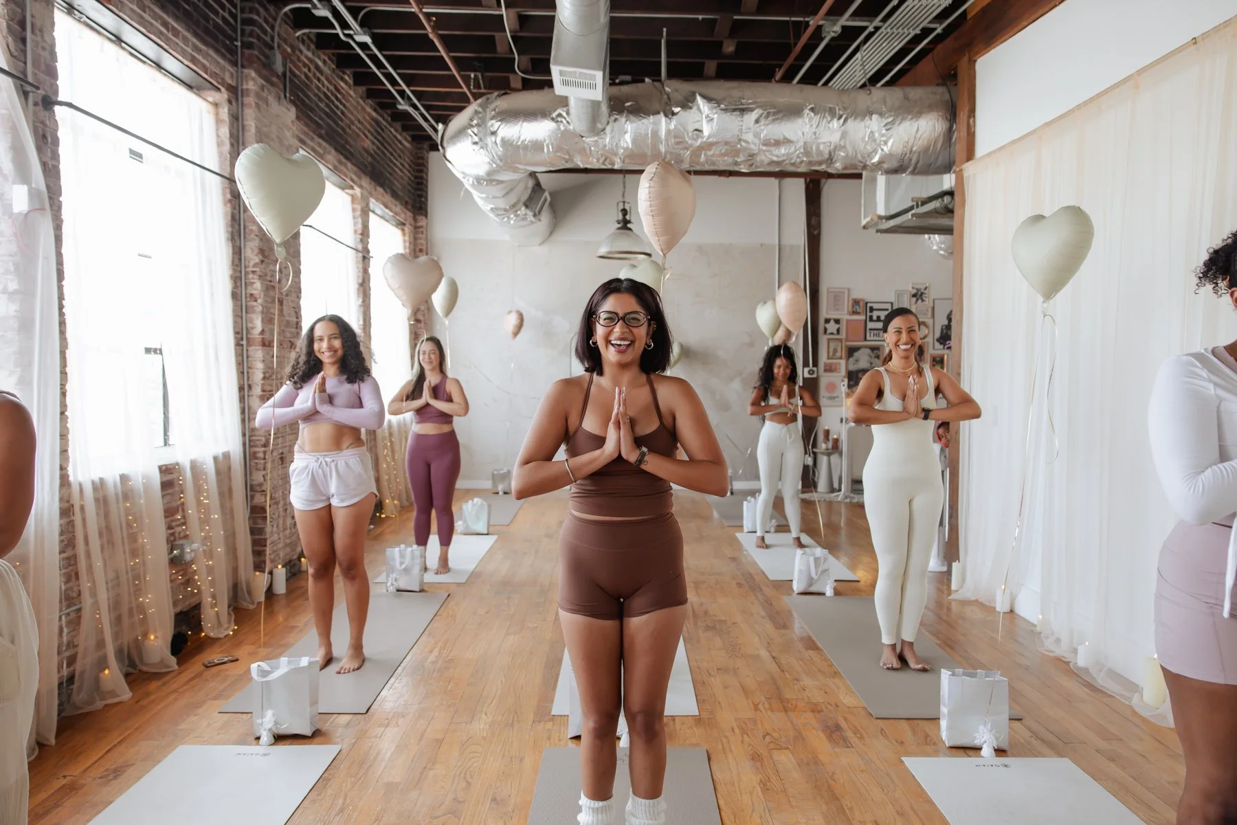 Women participating in a yoga or meditation class, standing on yoga mats, with some holding their hands in a prayer position, in a room decorated with balloons and fairy lights.