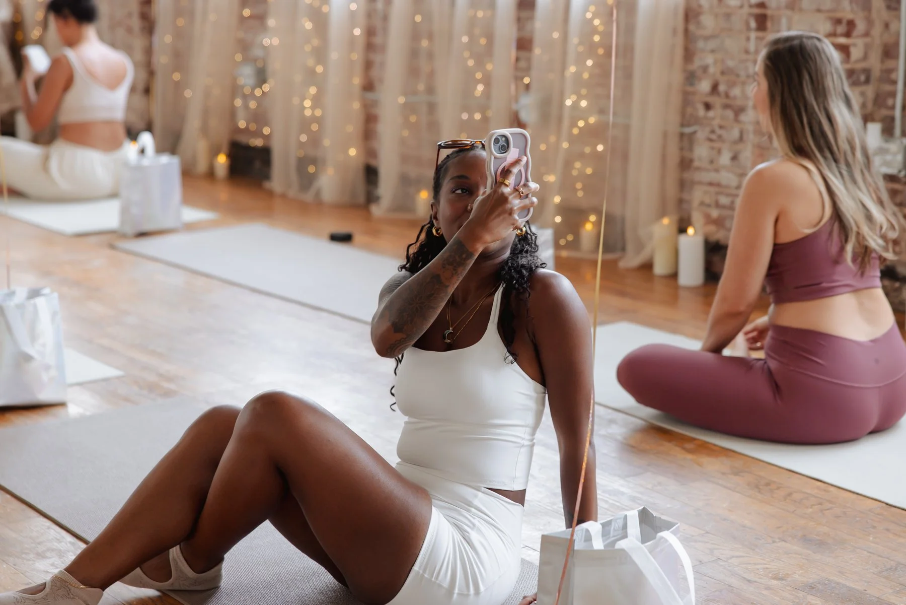 Woman sitting on yoga mat with phone in hand, practicing yoga in a cozy studio with exposed brick walls and string lights. Other women are practicing yoga in the background.