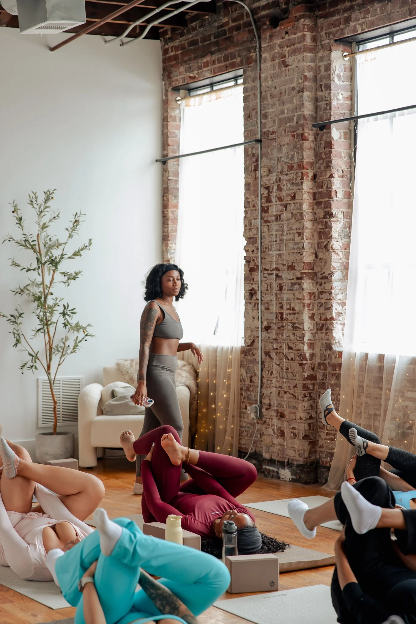 A group of people participating in a yoga class in a spacious studio with brick walls and large windows. Some are lying on mats practicing poses, while an instructor stands in the background overseeing the class.