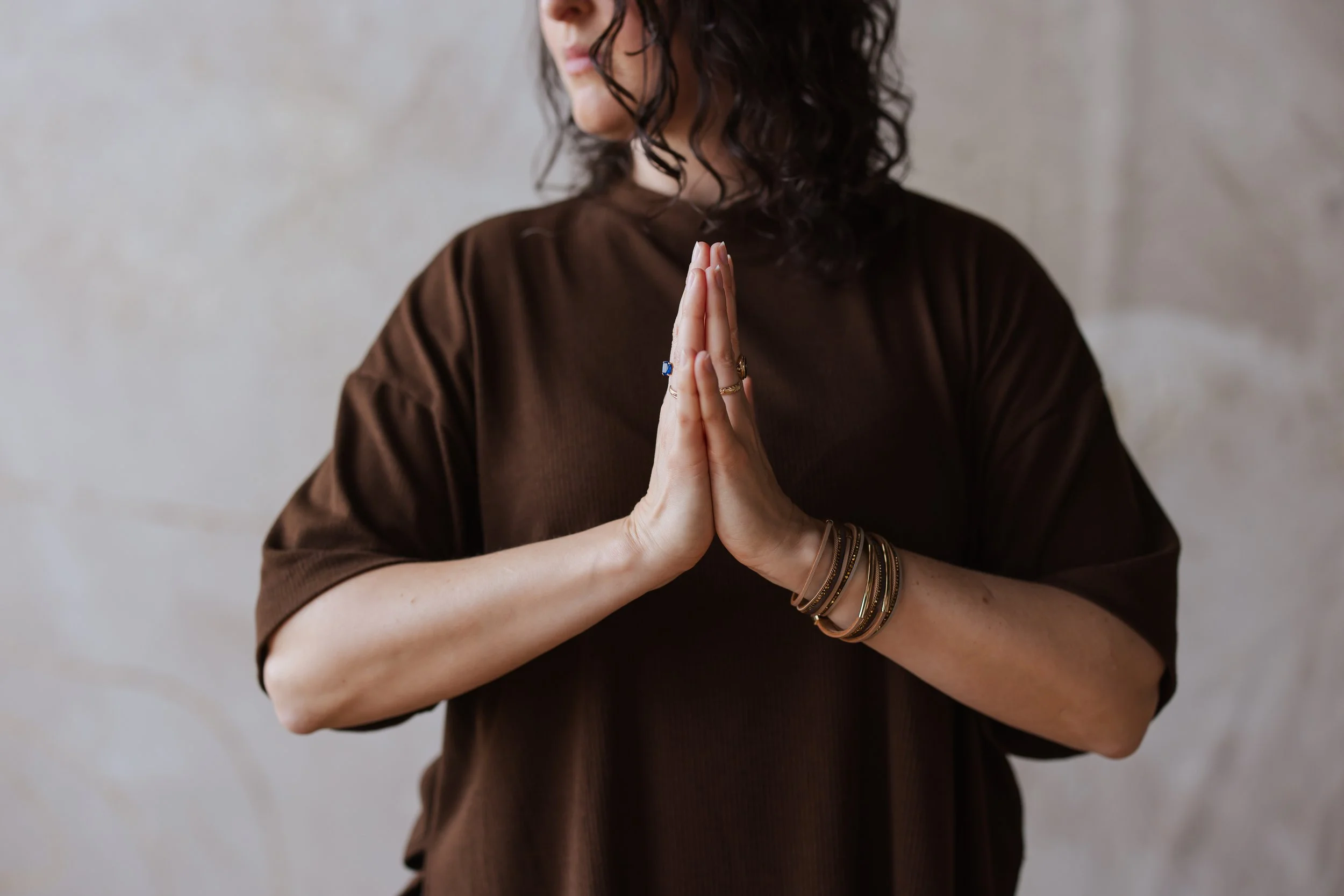 A woman with curly dark hair wearing a brown top, with her hands pressed together in a prayer-like gesture, adorned with rings and multiple bracelets, standing against a neutral background.