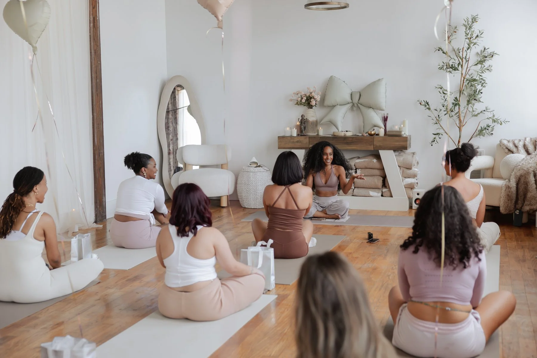 A woman leading a meditation or yoga session in a bright, cozy room with several women sitting cross-legged on mats, facing her. The room has white walls, a large mirror, a wooden shelf with candles, a decorative bow on the wall, and balloons, possib