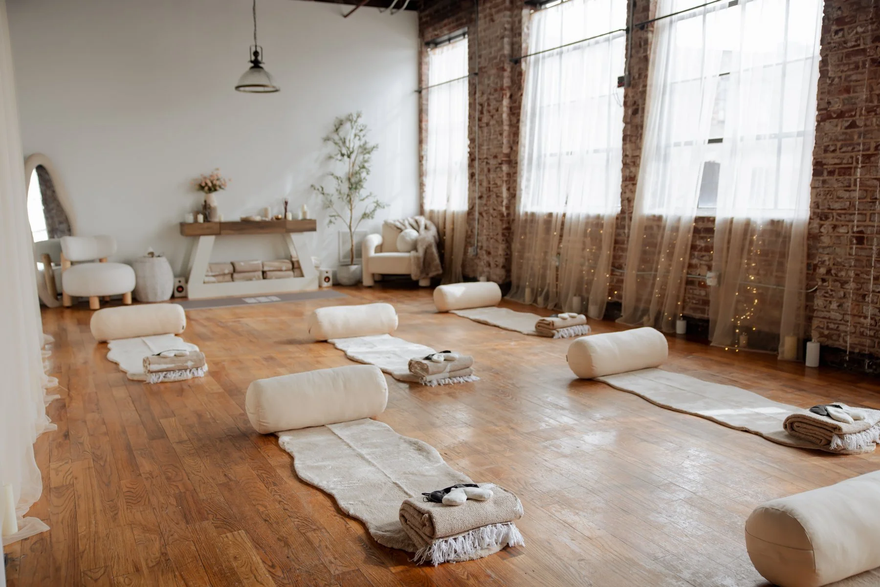 A spacious room set up for a yoga or meditation class with rolled towels, mats, and cushions arranged on a wooden floor, large windows with sheer curtains, exposed brick walls, and soft lighting.