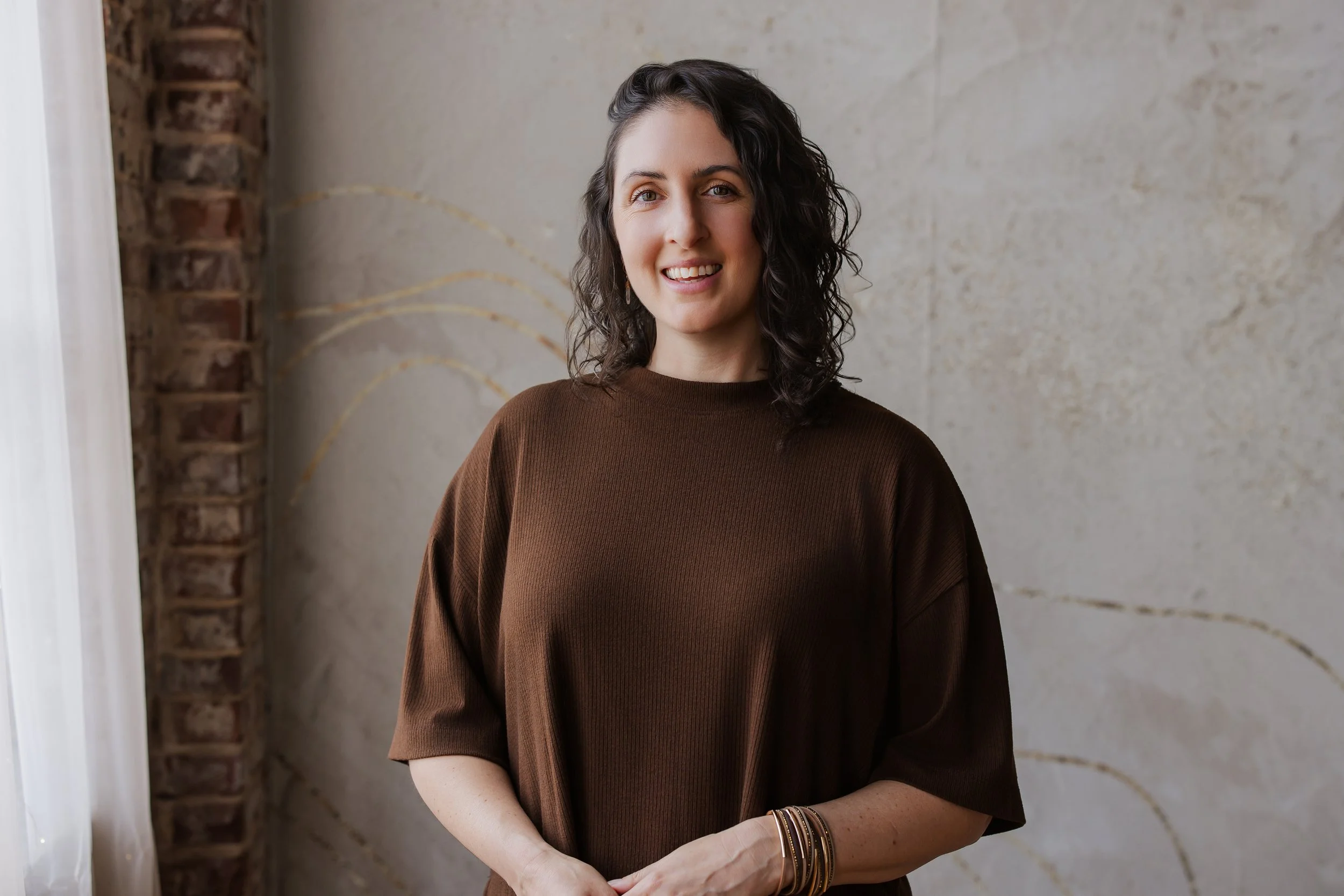 A woman with dark wavy hair smiling at the camera, wearing a brown shirt and gold bracelets, standing in front of a textured wall and a brick column.