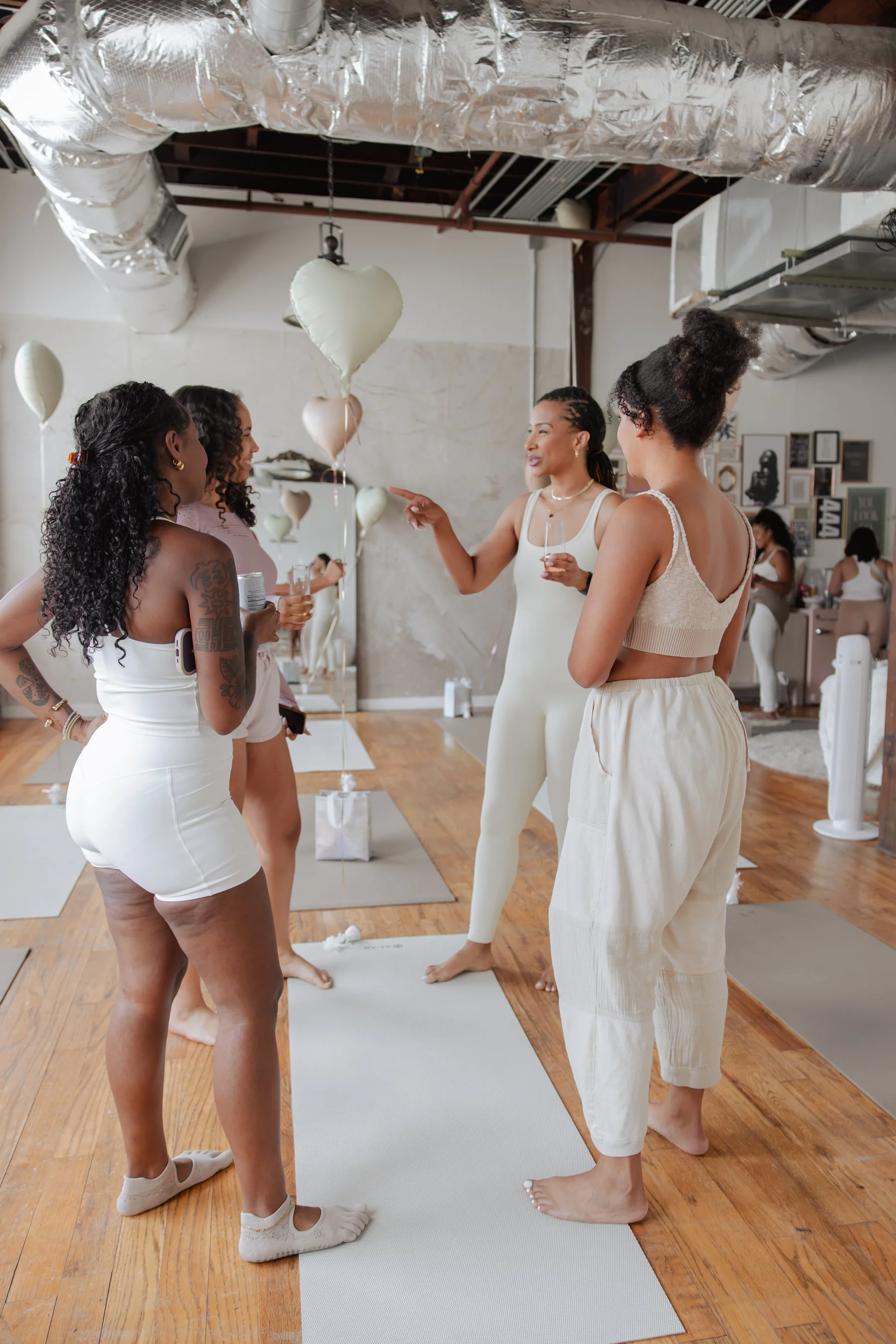 Group of women participating in a yoga or meditation class in a minimalist studio with hardwood floors, wearing light-colored clothing, and some holding drinks, standing on yoga mats while engaging with each other.