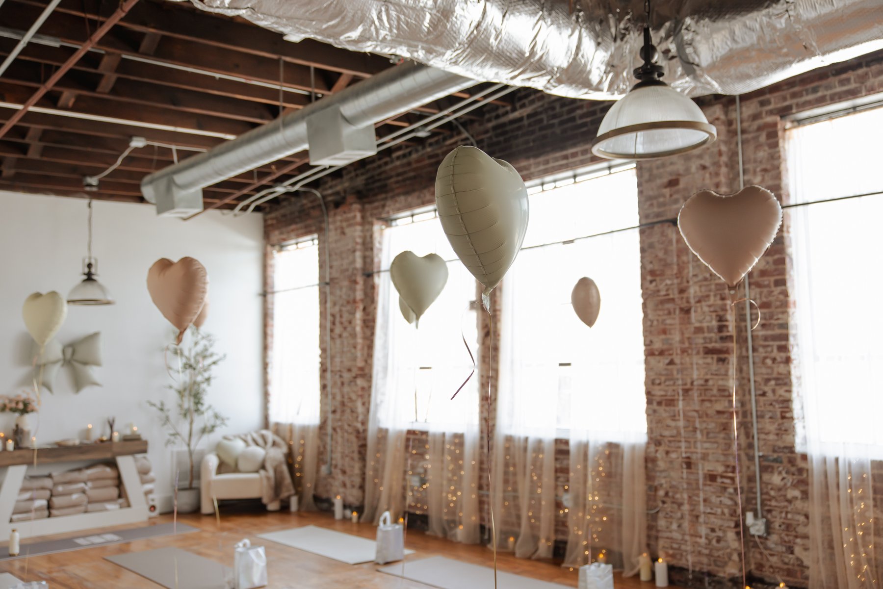 Interior of a rustic-style celebration space with brick walls and exposed ceiling beams, decorated with heart-shaped balloons in soft colors, fairy lights, and wrapped gifts on the floor.