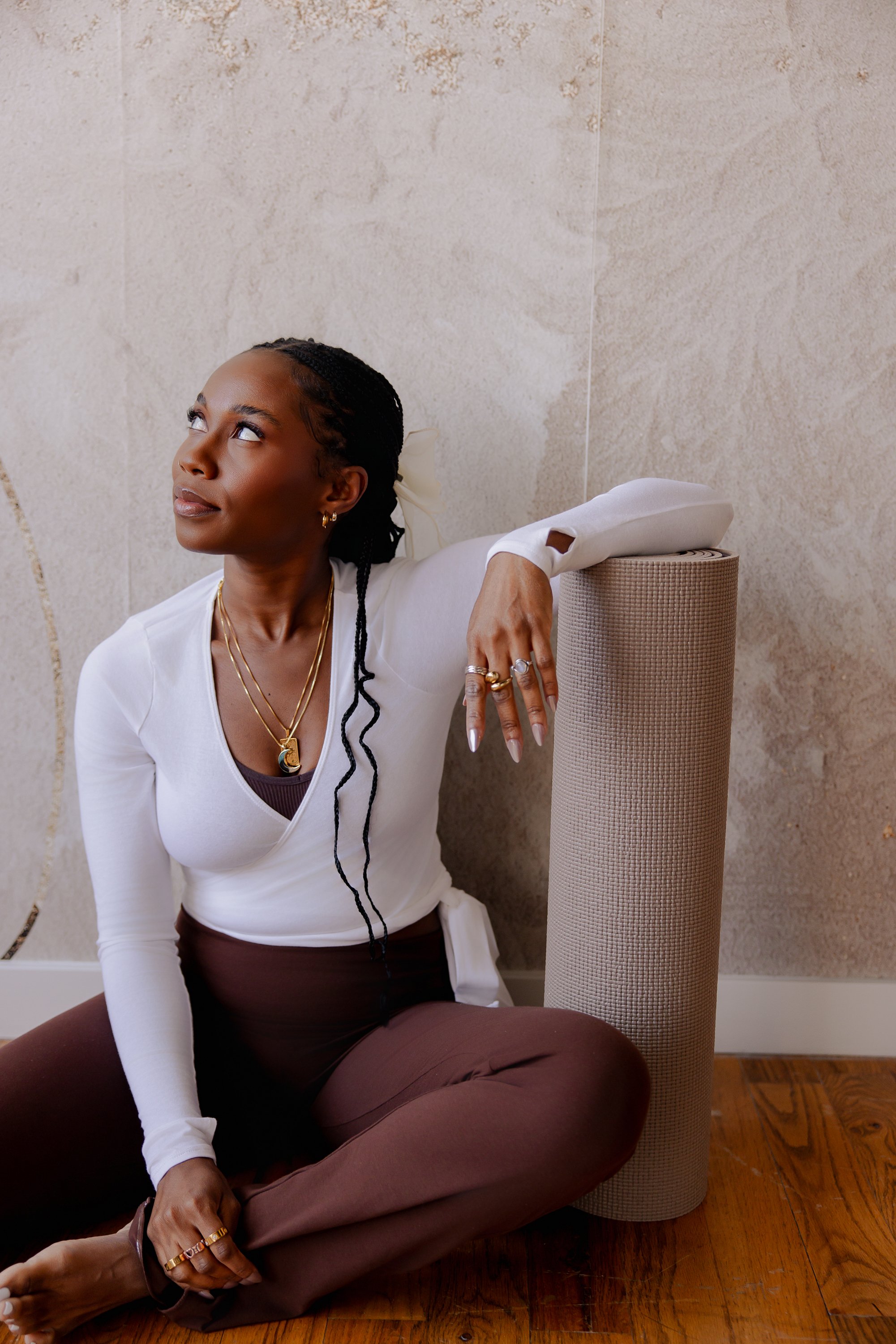 A woman sitting cross-legged on a wooden floor, leaning against a rolled-up yoga mat or foam roller, with a beige textured wall behind her. She has dark skin, braided hair, and is wearing a white long-sleeve top and brown pants, accessorized with layered necklaces and rings. She gazes slightly upward with a contemplative expression.