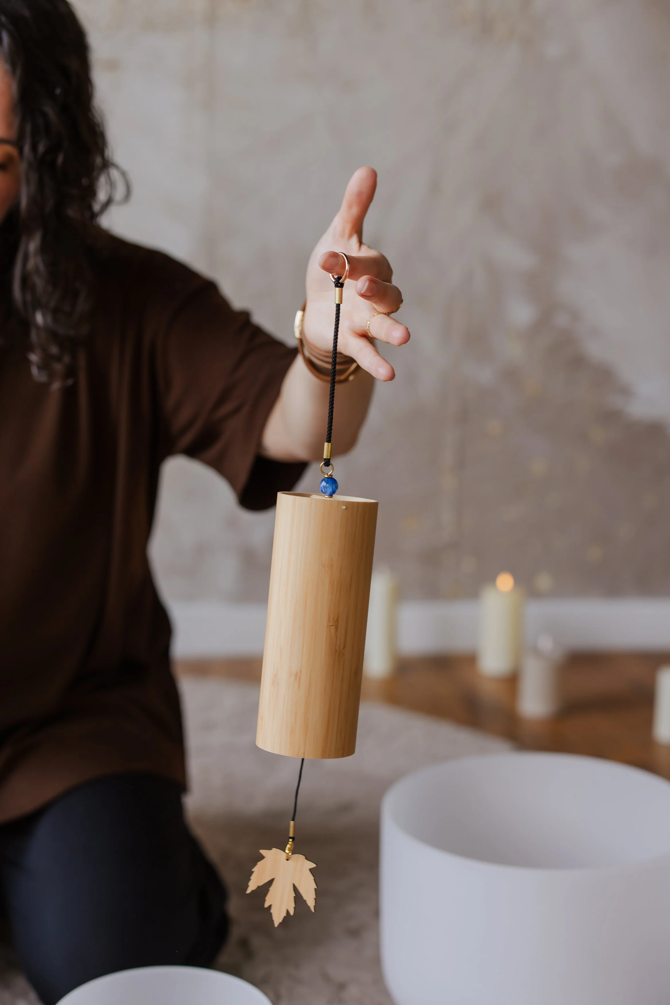 A person with curly hair wearing a brown shirt holds a string with a wooden leaf-shaped ornament attached, hanging from a wooden cylinder, in a room with candles and white chairs.