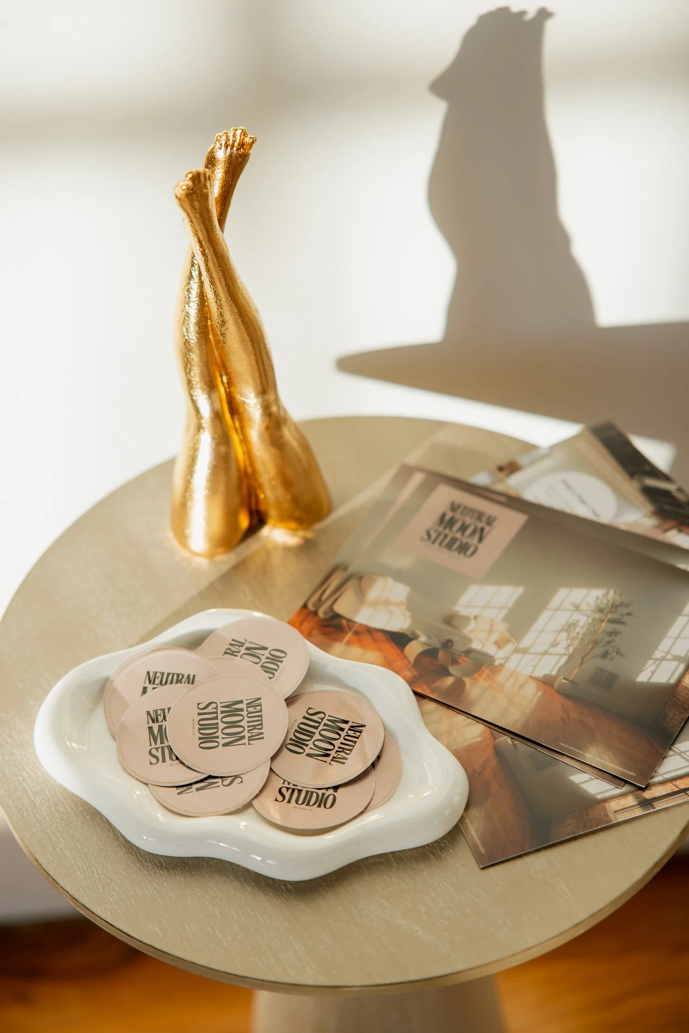 A round table with a gold hand sculpture, a white dish with beige tokens labeled 'NEUTRAL MOON STUDIOS', and magazines or brochures featuring interior design images.