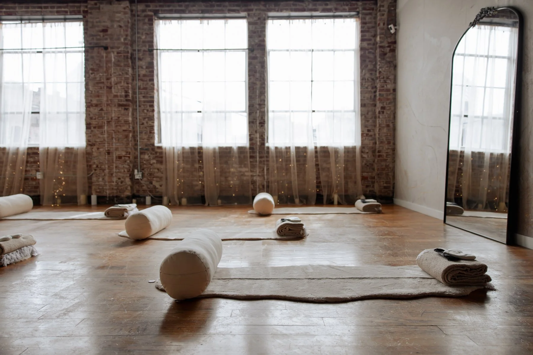 A yoga or meditation studio with wooden floors, large windows with sheer curtains, and brick walls. The room is set up with rolled-up towels and mats, suggesting a space for relaxation or yoga practice.