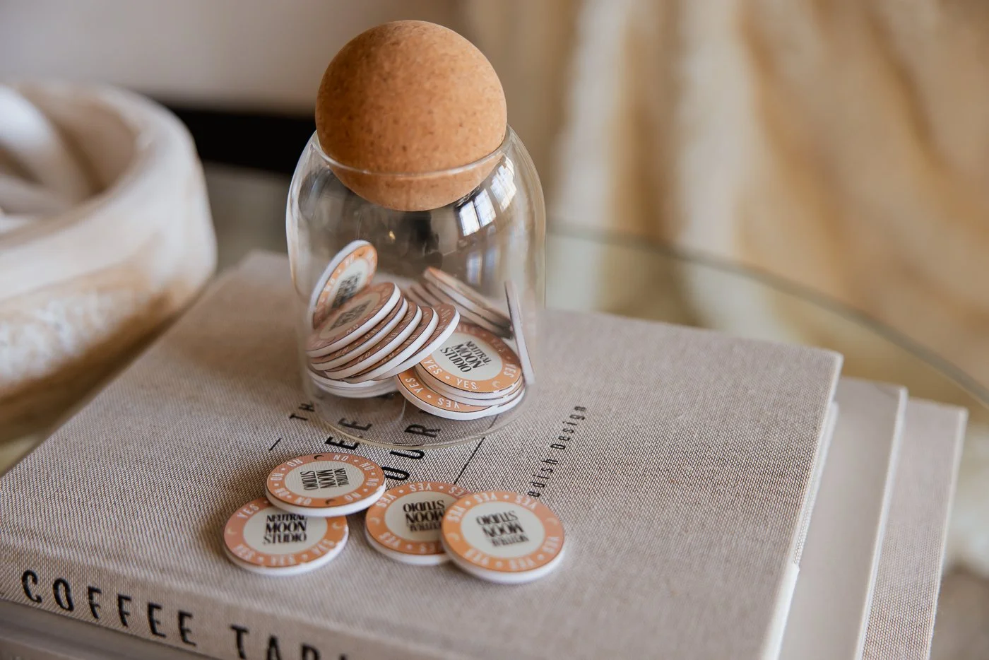 A glass jar with a cork lid filled with orange and white tokens labeled 'YES', 'NO', and 'MAYBE', placed on a stack of books and a beige cloth with text.
