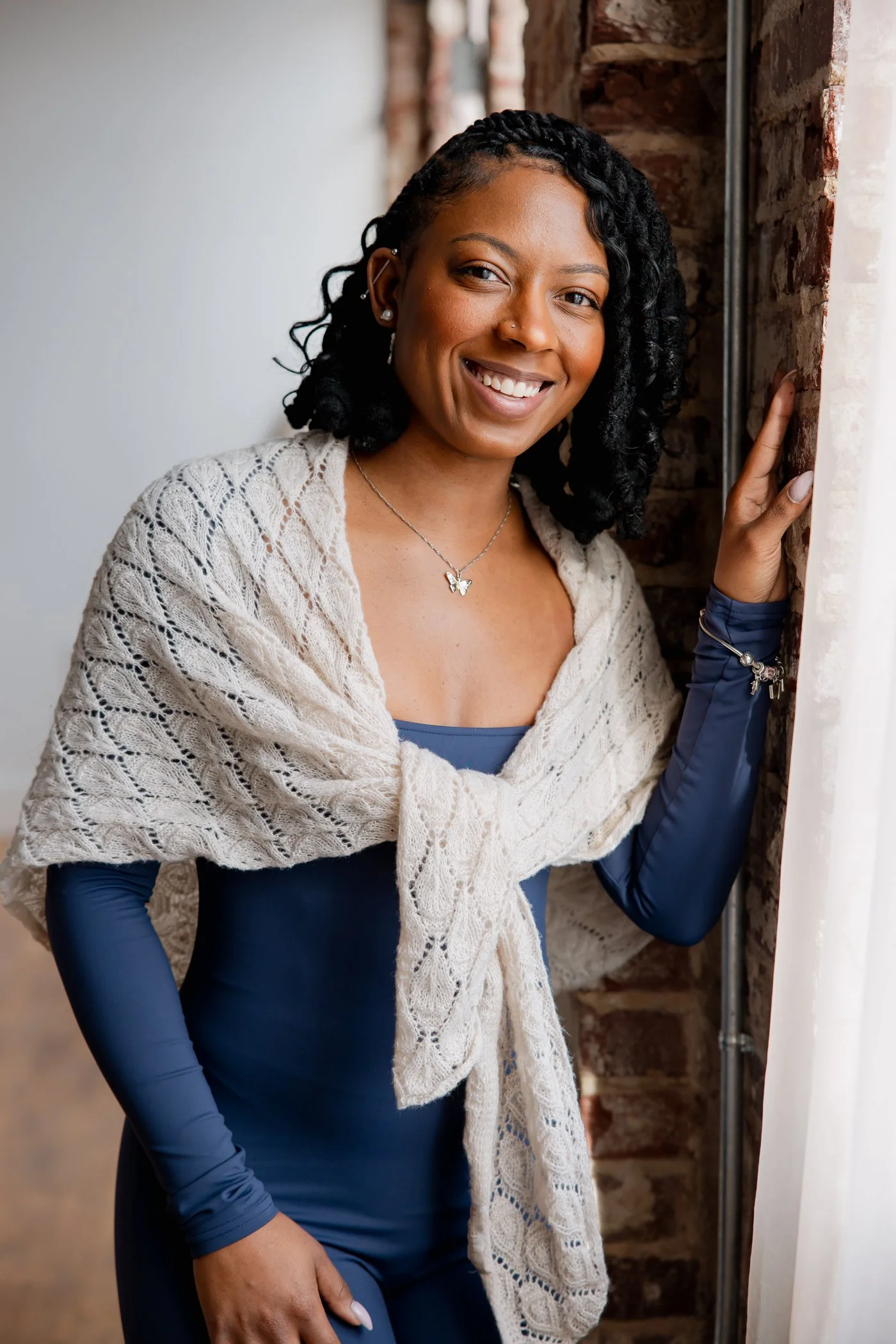 Smiling woman with dark hair in braids, wearing a navy dress, cream knitted shawl, and jewelry, standing next to a brick wall and window.