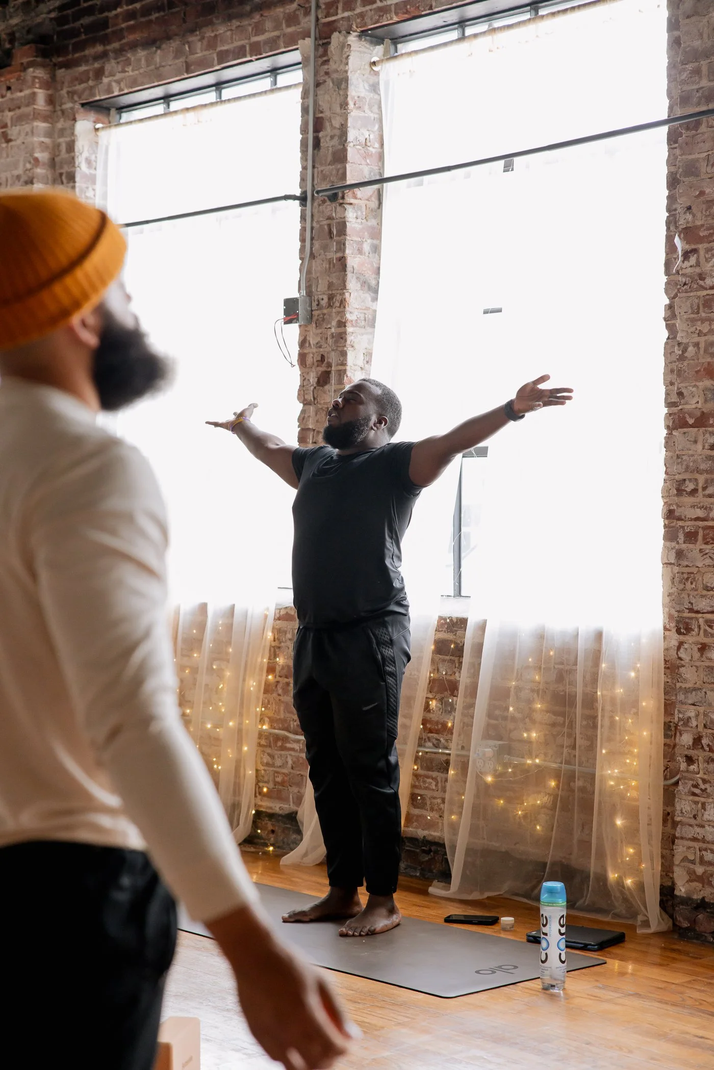 A man in black athletic wear standing on a yoga mat with arms outstretched, leading a yoga or meditation class in a studio with exposed brick walls and large windows. Another person is partially visible in the foreground, wearing a white top and yellow beanie.