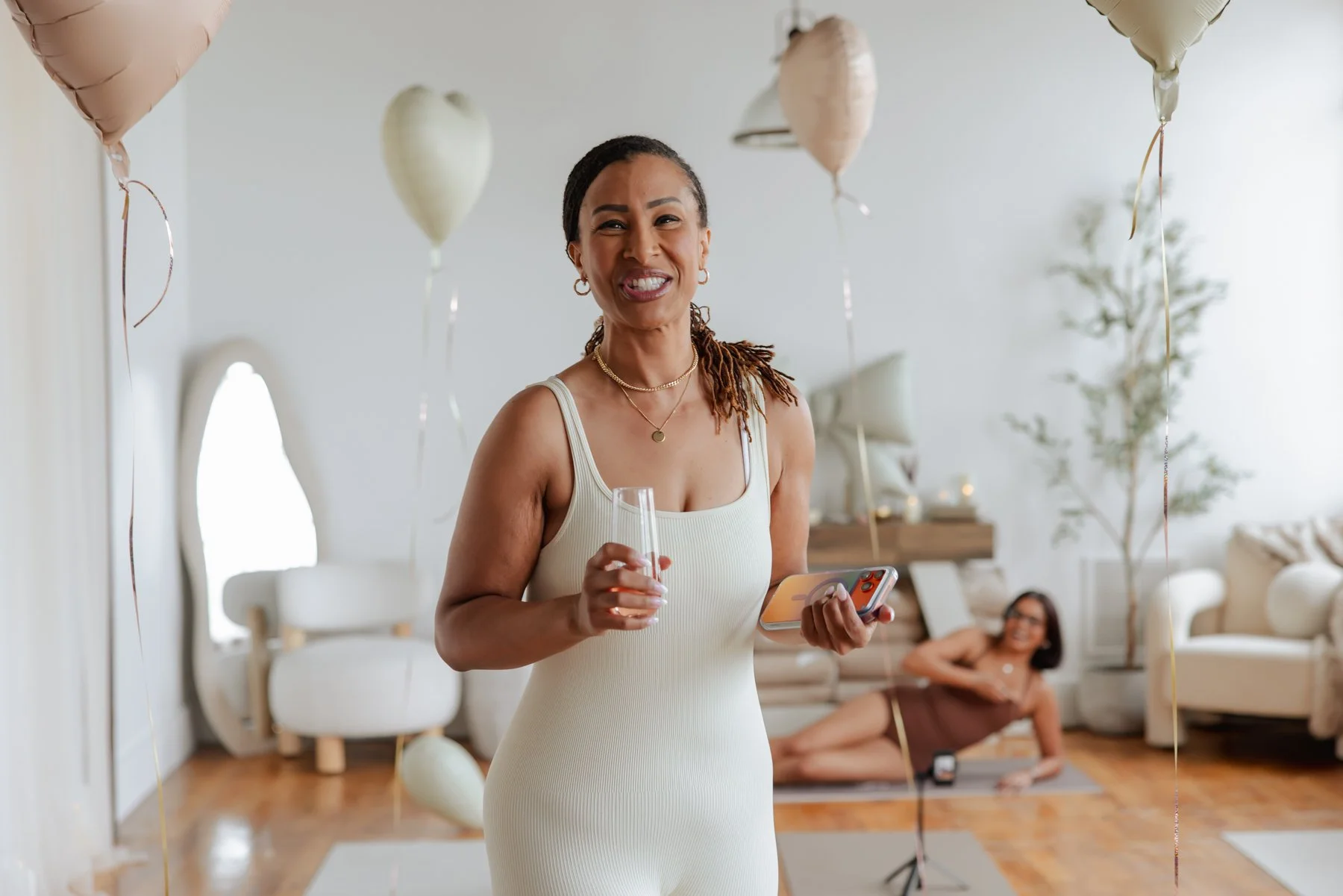A woman with dark hair styled in cornrows and wearing a cream-colored sleeveless dress is smiling and holding a glass of wine and a cellphone at a celebration. In the background, another woman with glasses, wearing a brown dress, is sitting on the fl