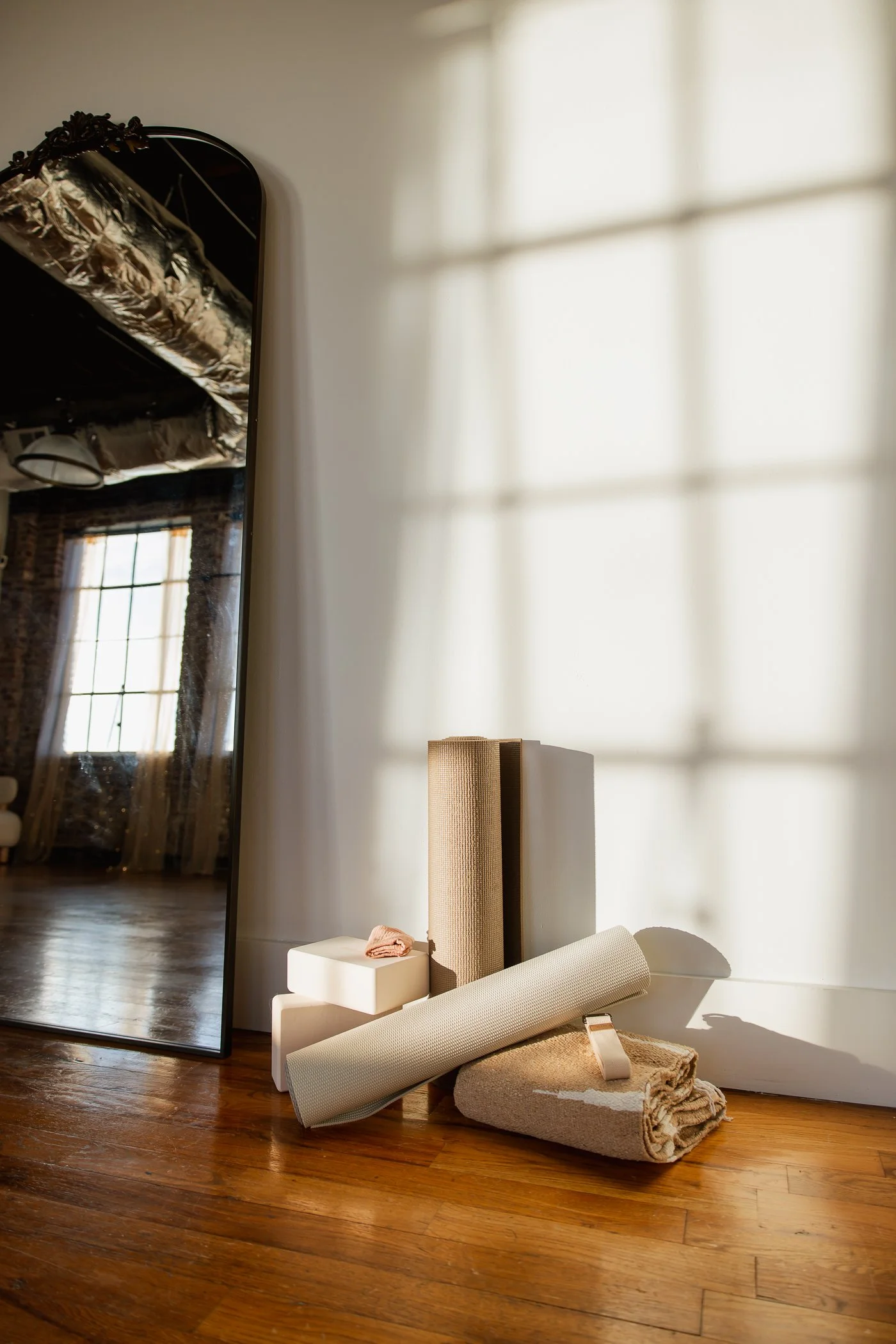 Stacked yoga mats and towels next to a large mirror in a sunlit room with wooden floors.