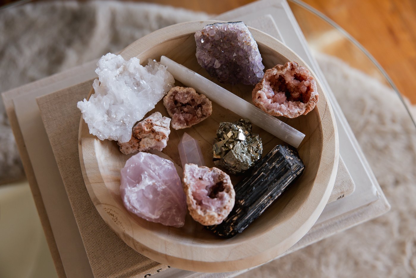 A wooden bowl filled with various raw crystals and minerals, including quartz, rose quartz, hematite, and amethyst, on a fabric-covered surface.