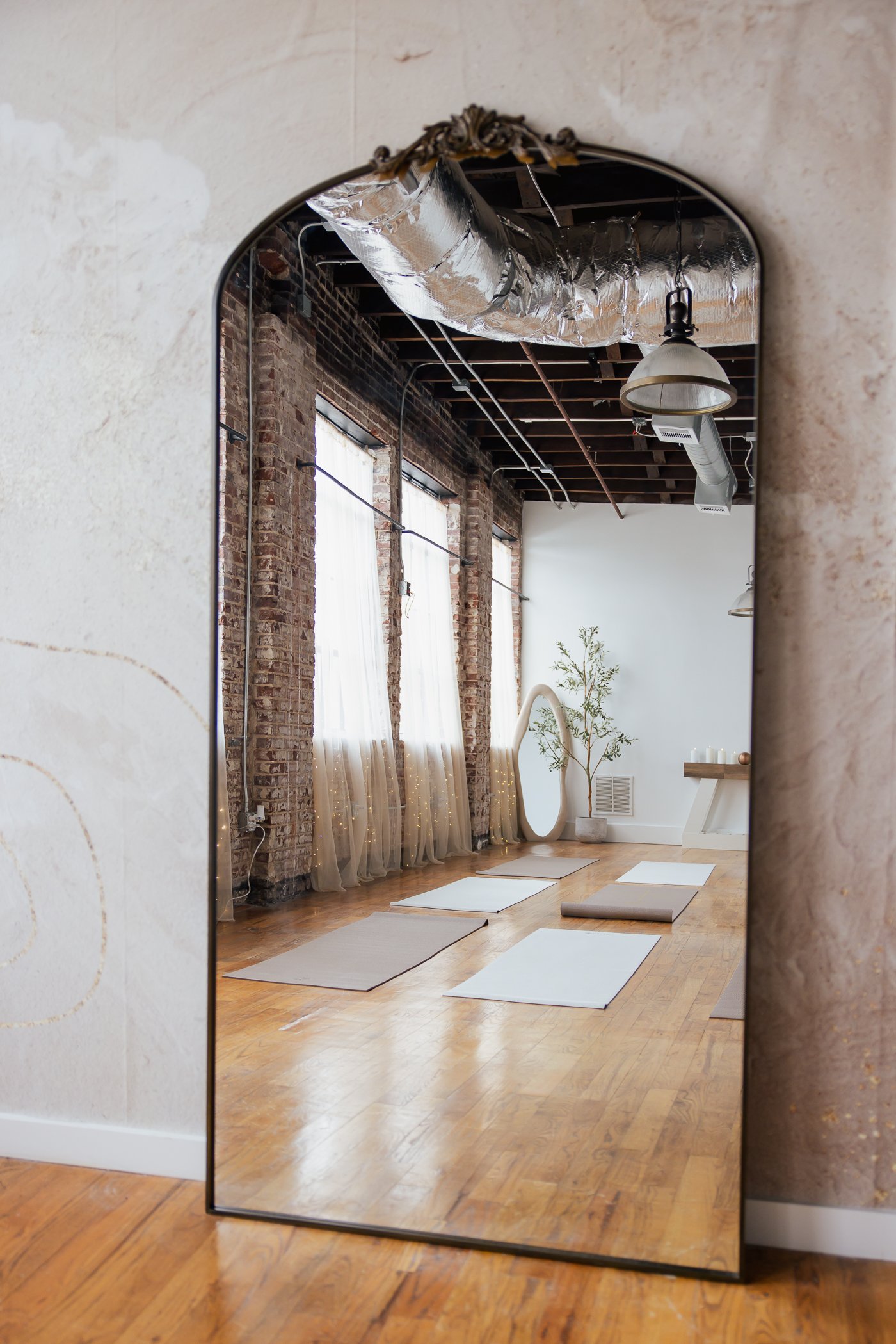 Empty yoga studio with mats on wooden floor, large mirror, exposed brick walls, tall windows with sheer curtains, and a potted plant.