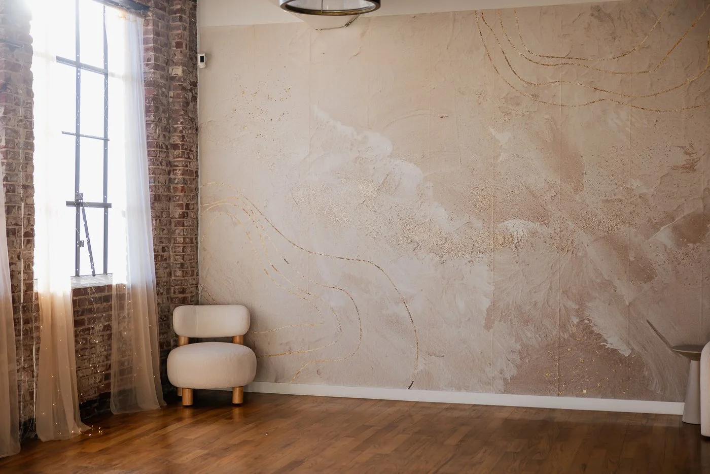 A spacious room with a brick wall and a large window with sheer curtains. There is a white cushioned chair with wooden legs on a wooden floor, against a marble-patterned wall.