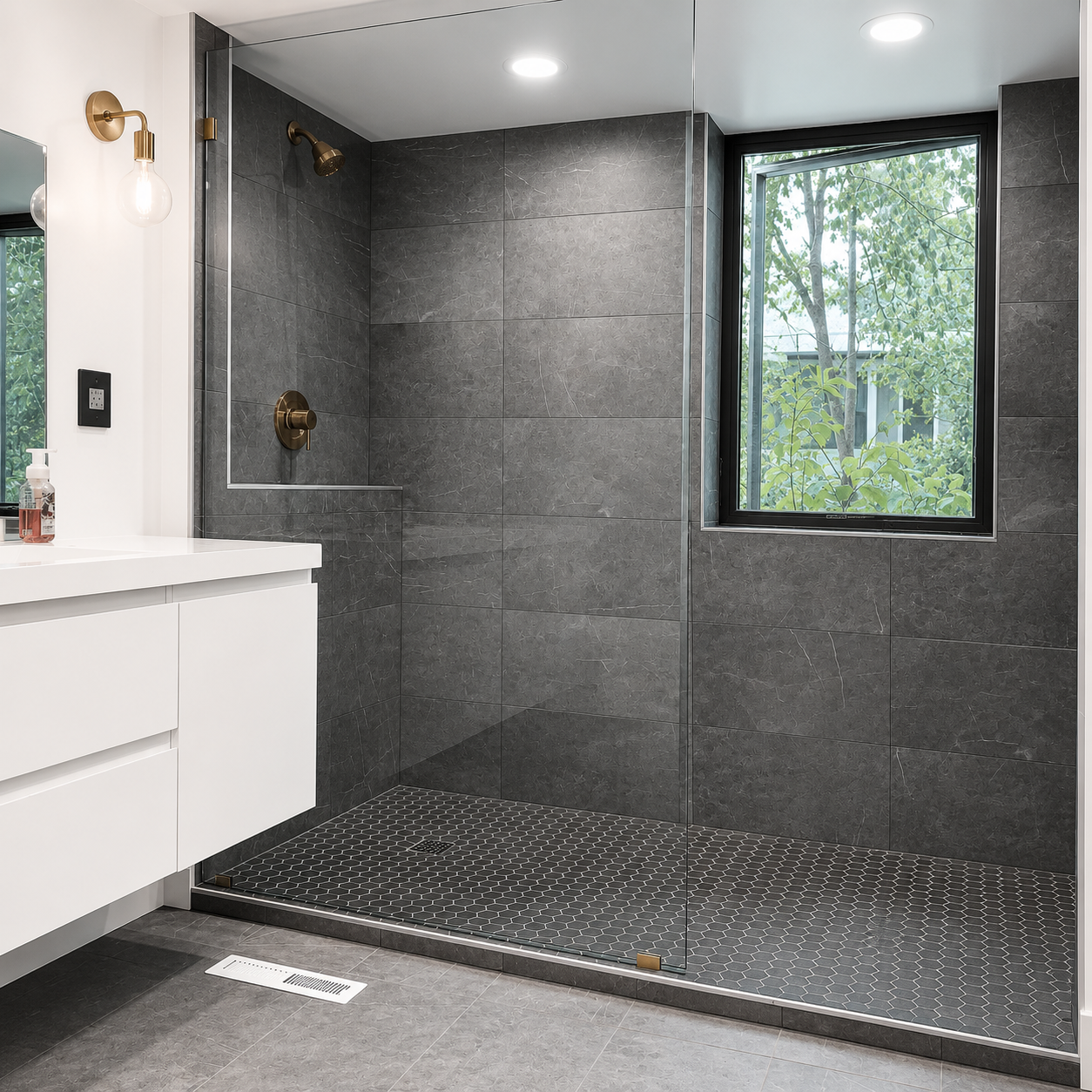 Modern bathroom with a walk-in shower, dark gray tiles, a window showing greenery outside, a white vanity with a mirror, and brass fixtures.