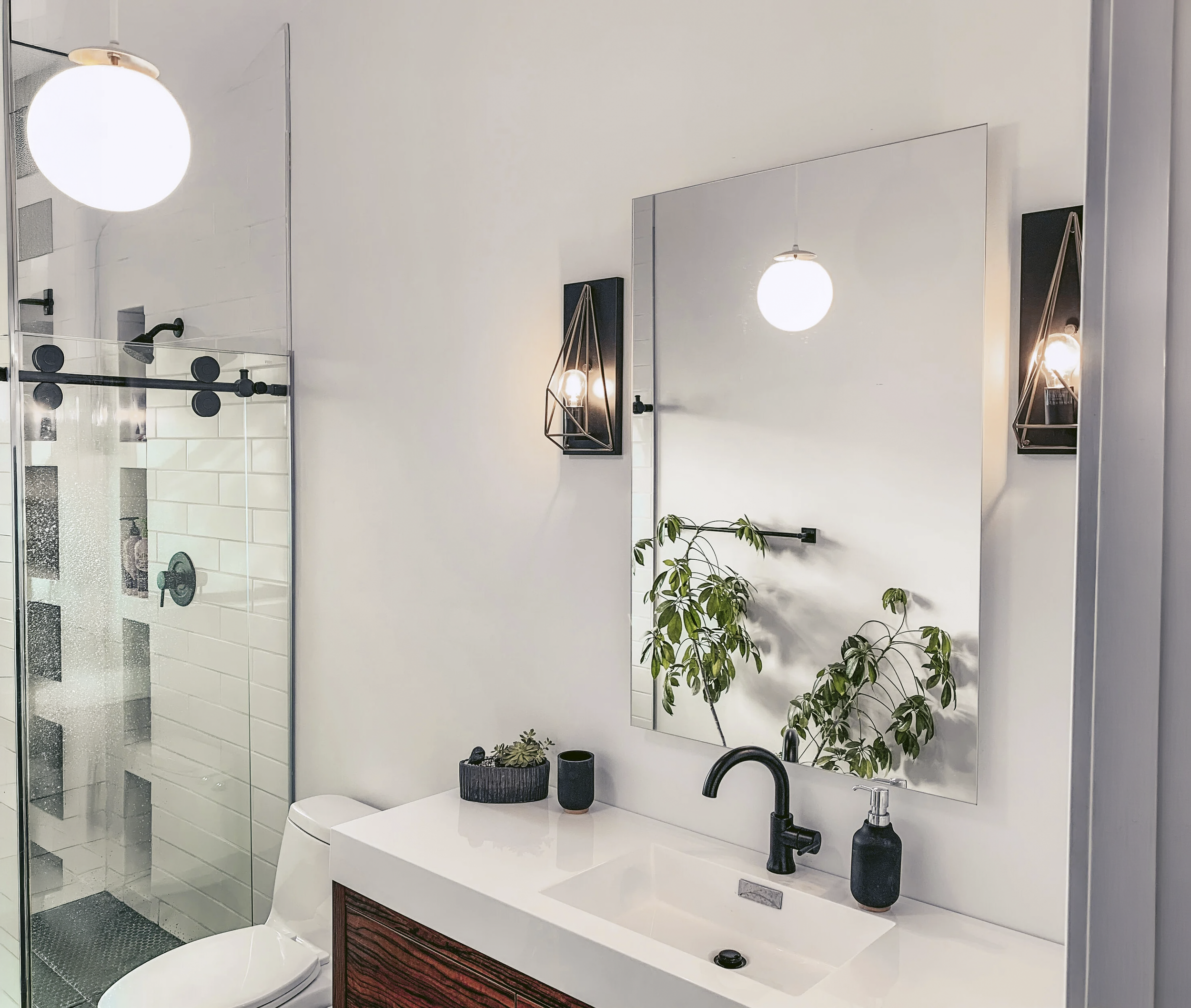 Modern bathroom with white walls, a large mirror, black fixtures, a wooden vanity, potted plants, and a glass-enclosed shower with white subway tiles.