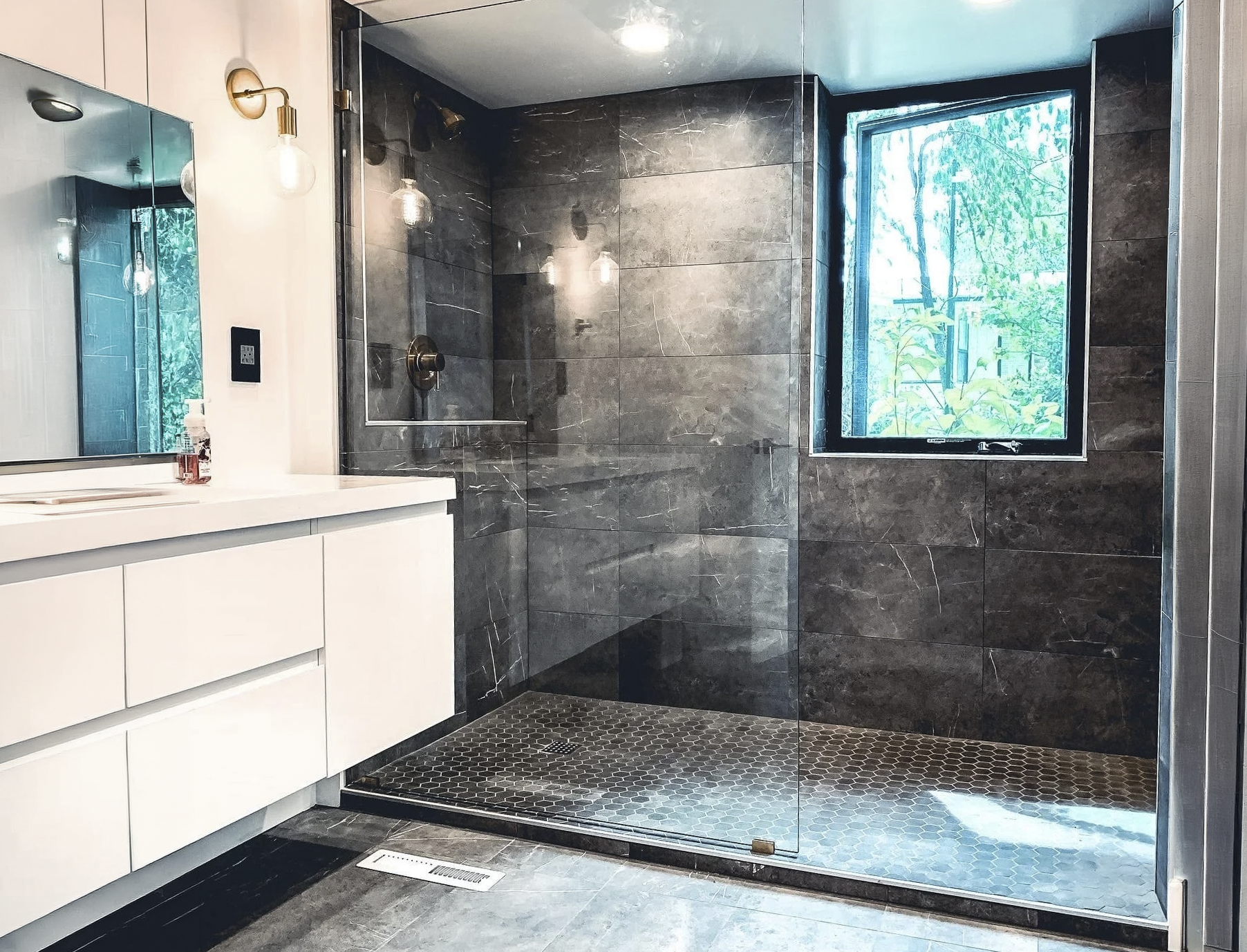 Modern bathroom with a walk-in shower enclosed by glass, dark gray tiled walls, a black honeycomb-patterned floor, a window showing green trees outside, and a white vanity with a mirror and light fixtures.