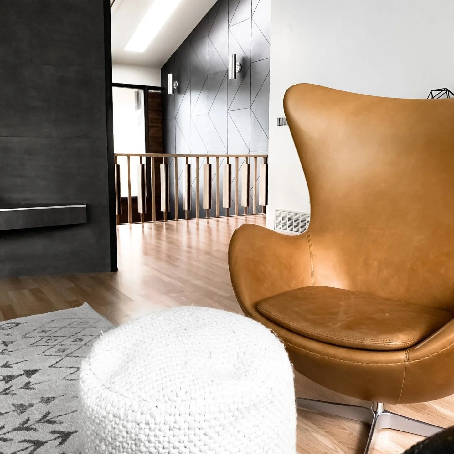 Modern interior living space with a tan leather lounge chair, a white knitted pouf, a patterned rug, wooden floors, a wooden railing, and geometric wall decor.