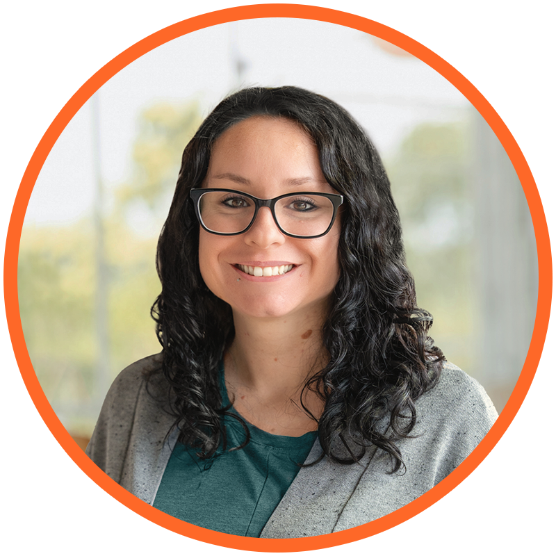 A woman with curly black hair, glasses, and a grey blazer smiling in a well-lit room with windows and greenery in the background.