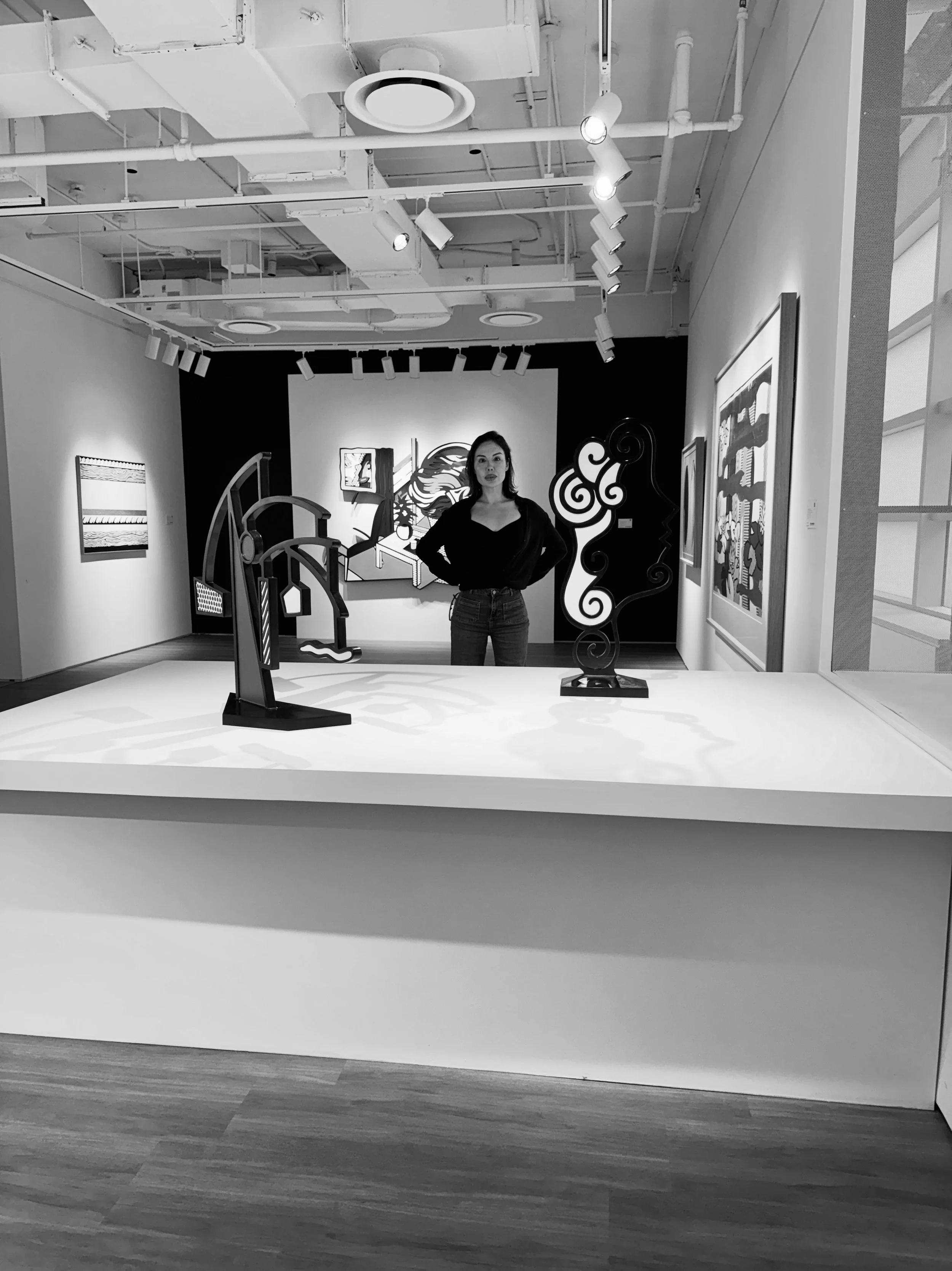 Black and white photo of an art gallery with paintings on the walls and a woman standing in the center. The gallery has a high ceiling with visible pipes and track lighting.