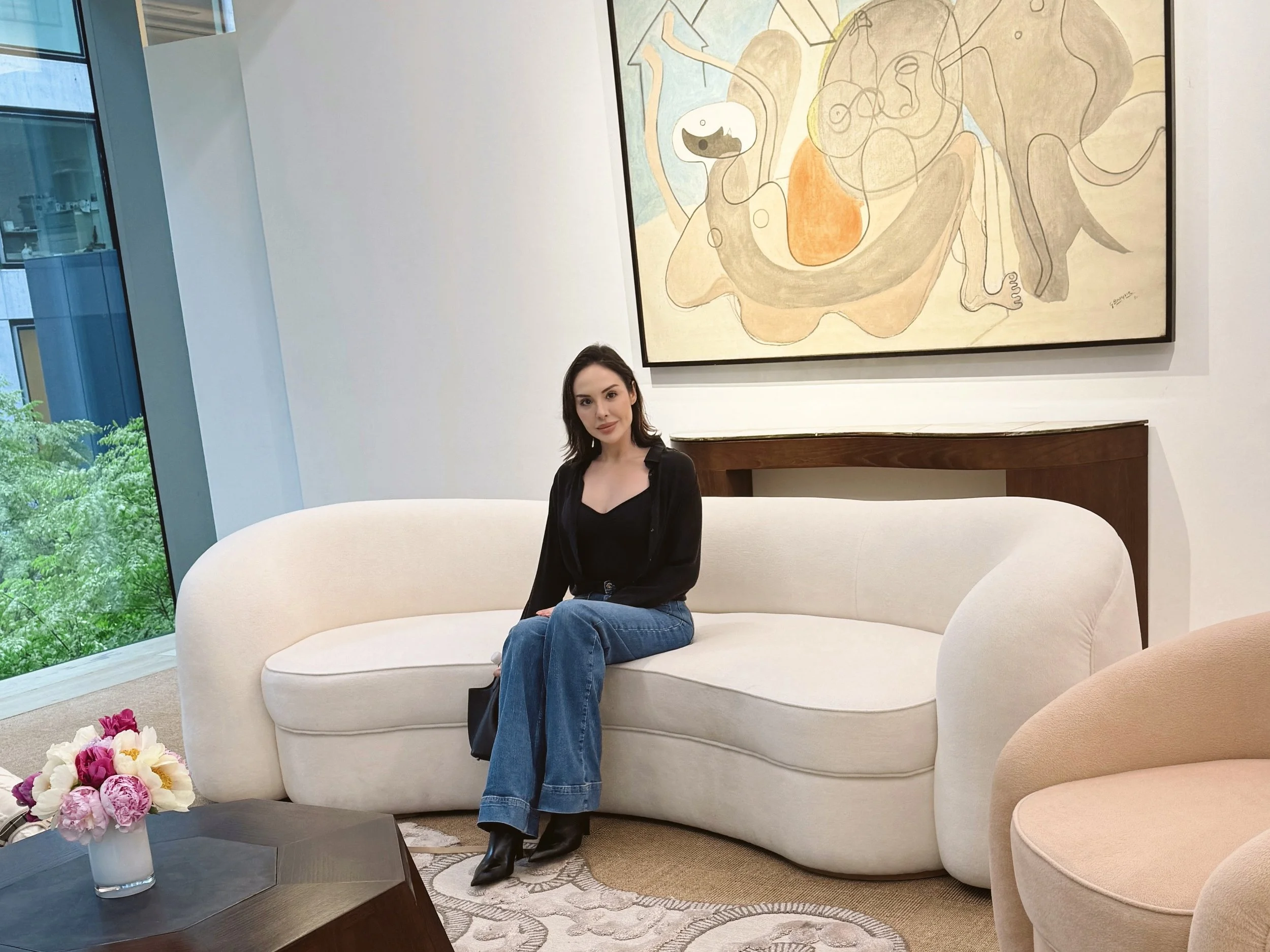A woman with shoulder-length dark hair wearing a black top, blue jeans, and black heeled boots sitting on a white curved sofa in a modern, brightly-lit room. Behind her, there is a large abstract painting with soft beige, gray, and orange tones. To her left, there is a glass-and-metal coffee table with a white vase of pink and white flowers. On the left side of the image, there is a large window showing a view of green trees outside.