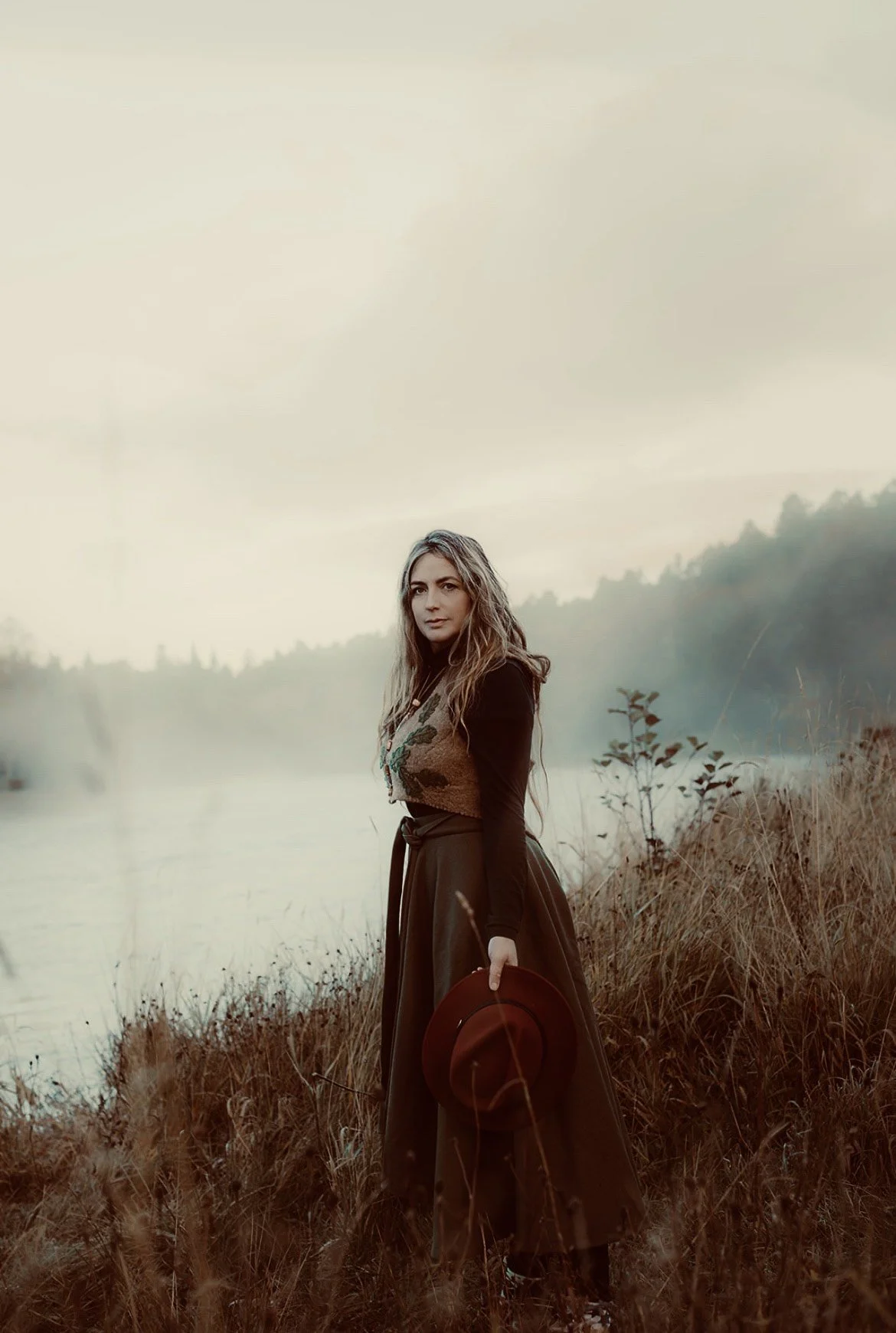 A woman with long hair standing outdoors near a body of water, holding a brown hat in one hand, surrounded by tall grasses and with a misty landscape in the background.